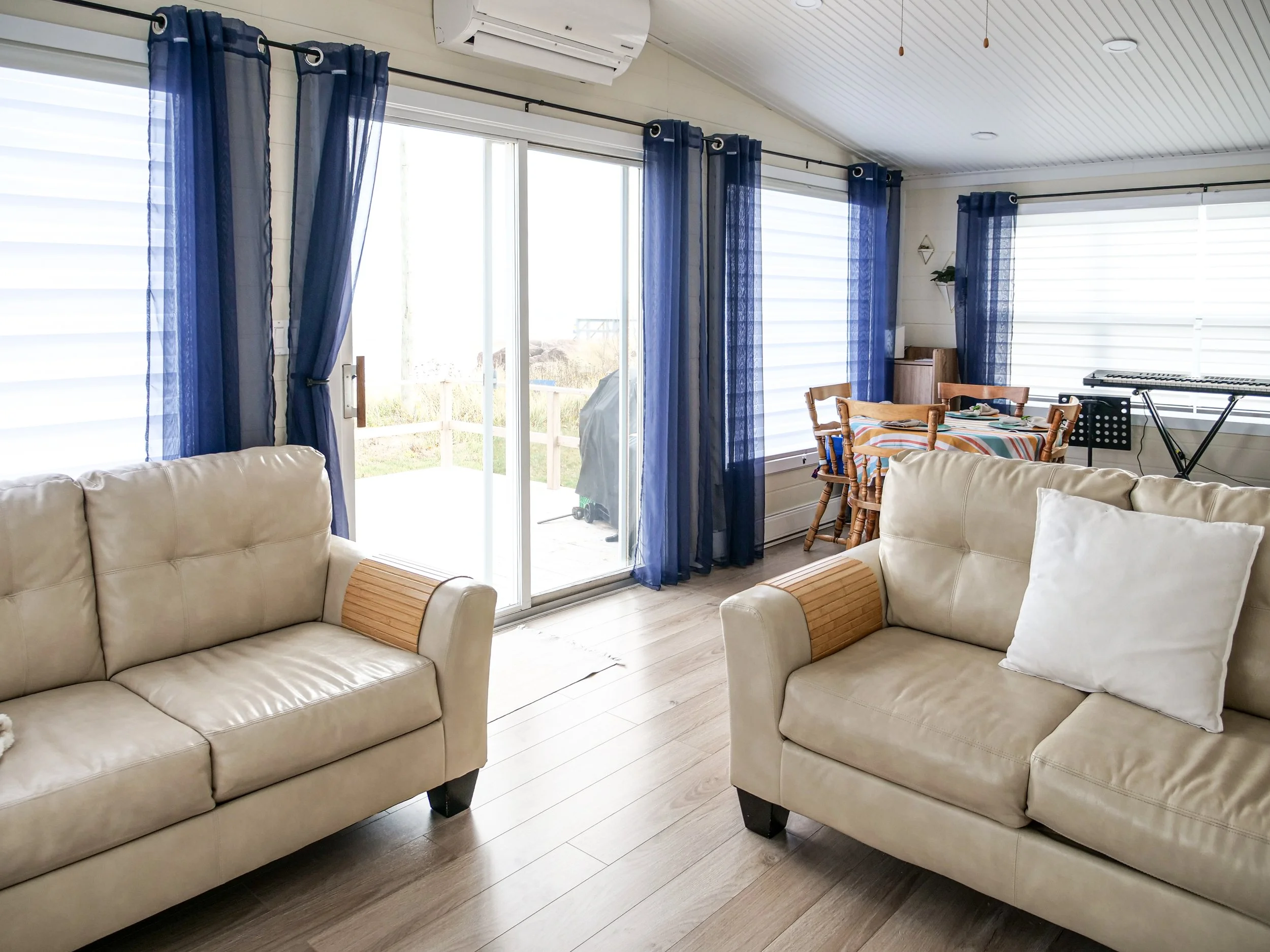 Living room with beige sofas, blue curtains, a dining table with chairs, a keyboard, and sliding glass doors leading outside
