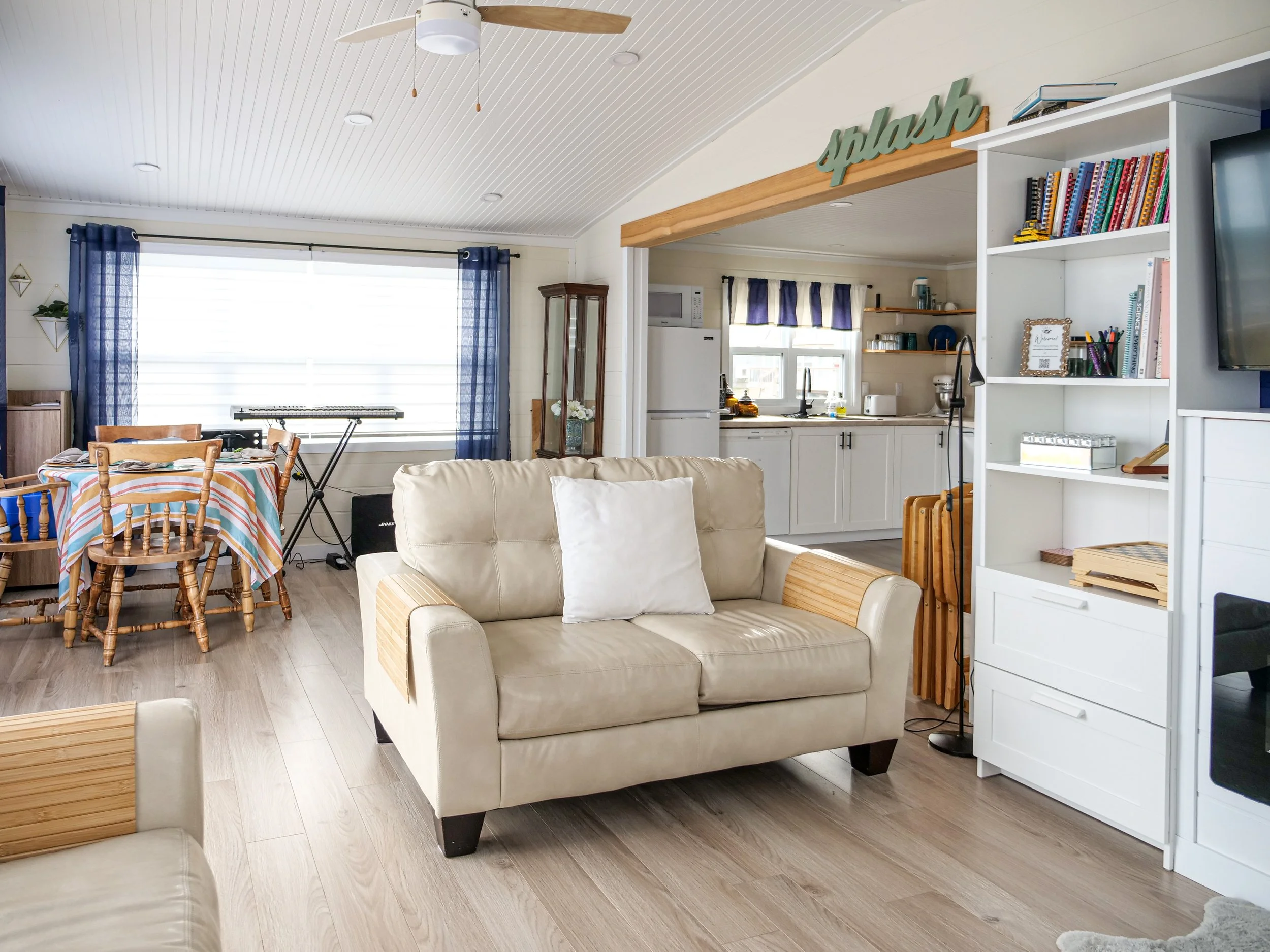 Living room and kitchen area with a beige sofa, a dining table with chairs, bookshelves, and large windows with blue curtains.