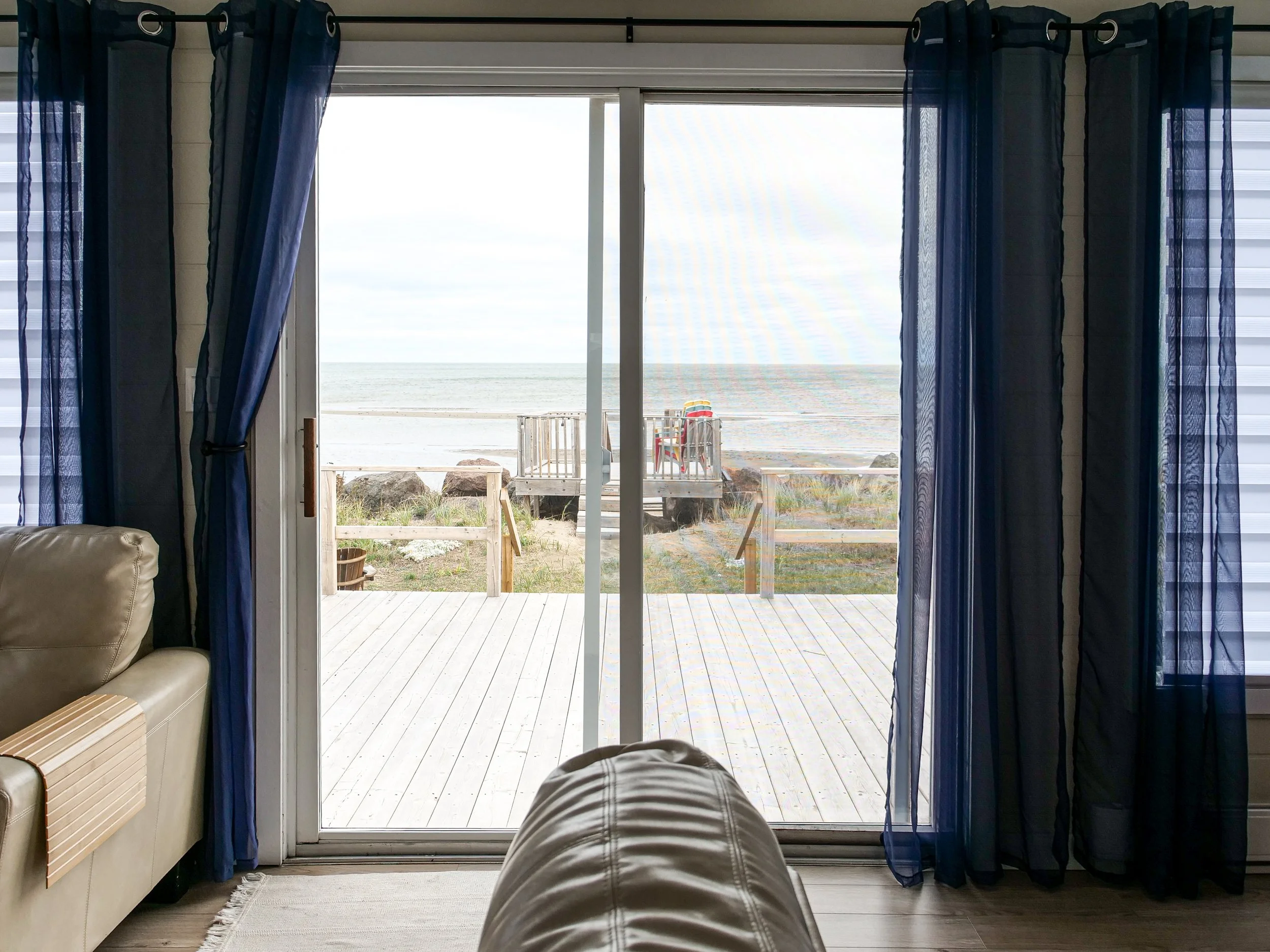View of a beach from inside a house looking through sliding glass doors, with a sandy shore, rocks, and a wooden deck outside.