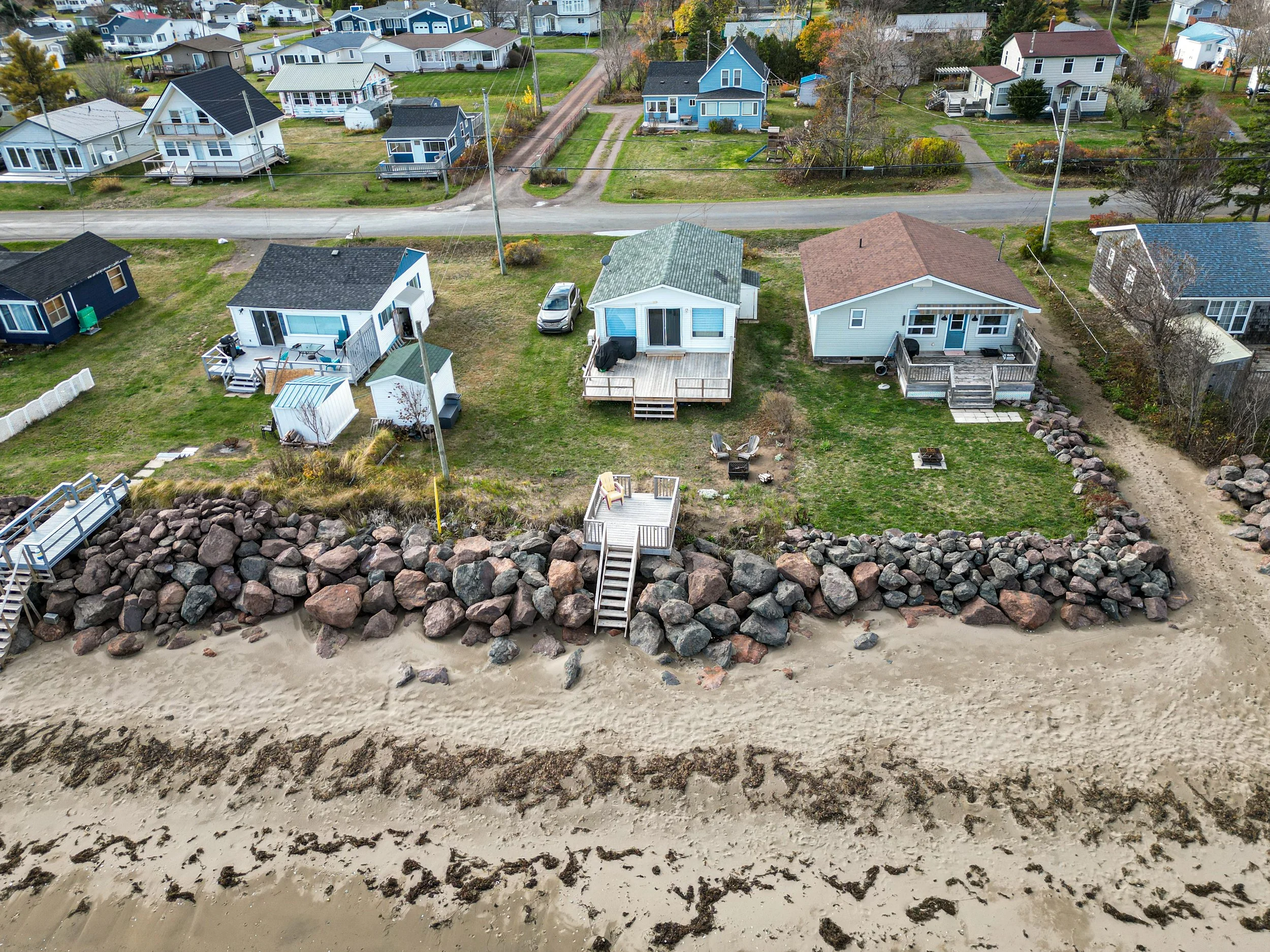 Aerial view of a row of houses on a beach, with a stone wall separating the sand from the backyards, which have grass, decks, outdoor furniture, and a few trees.