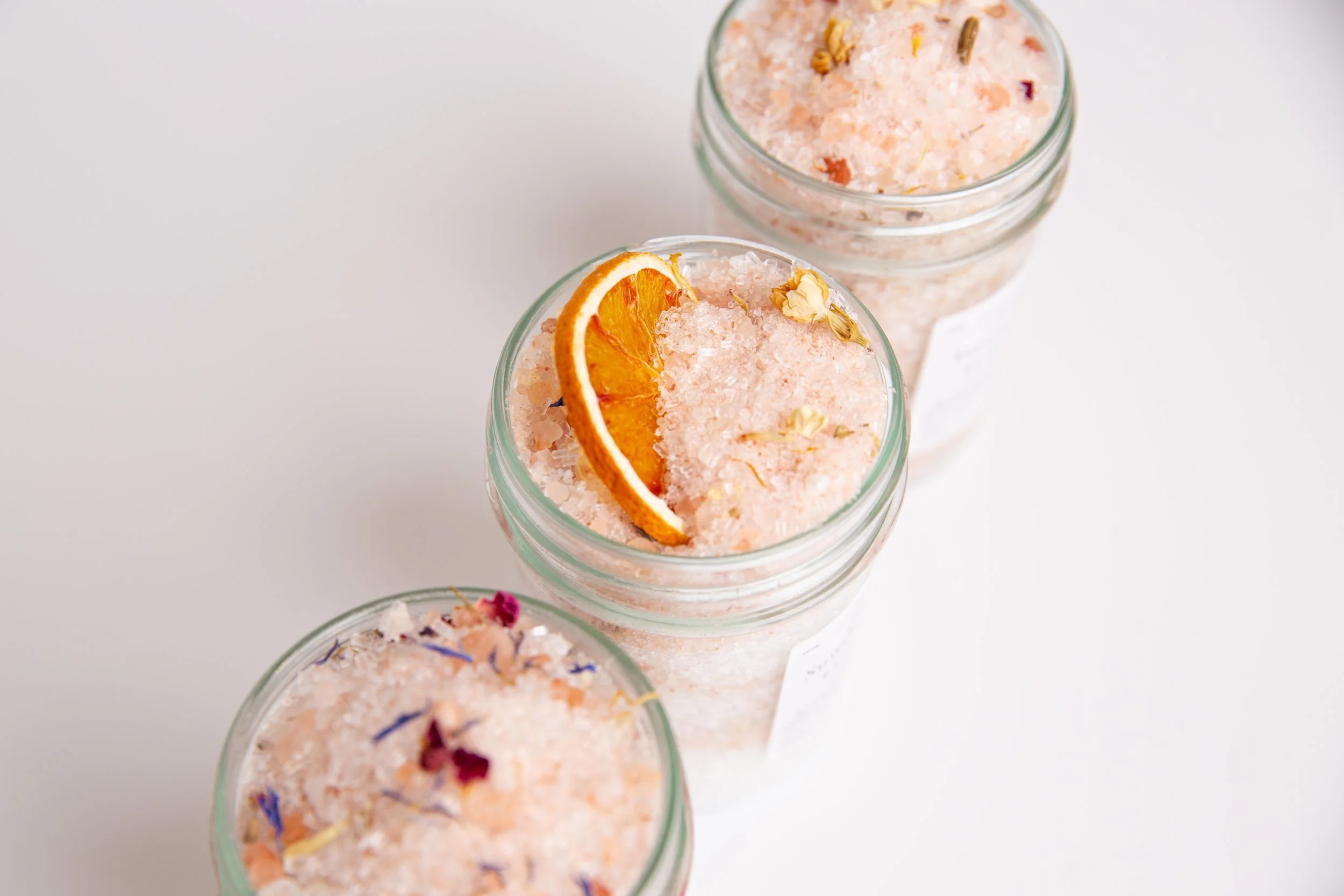 Close-up of three glass jars filled with pink Himalayan salt, garnished with dried orange slices and flower petals, on a white surface.