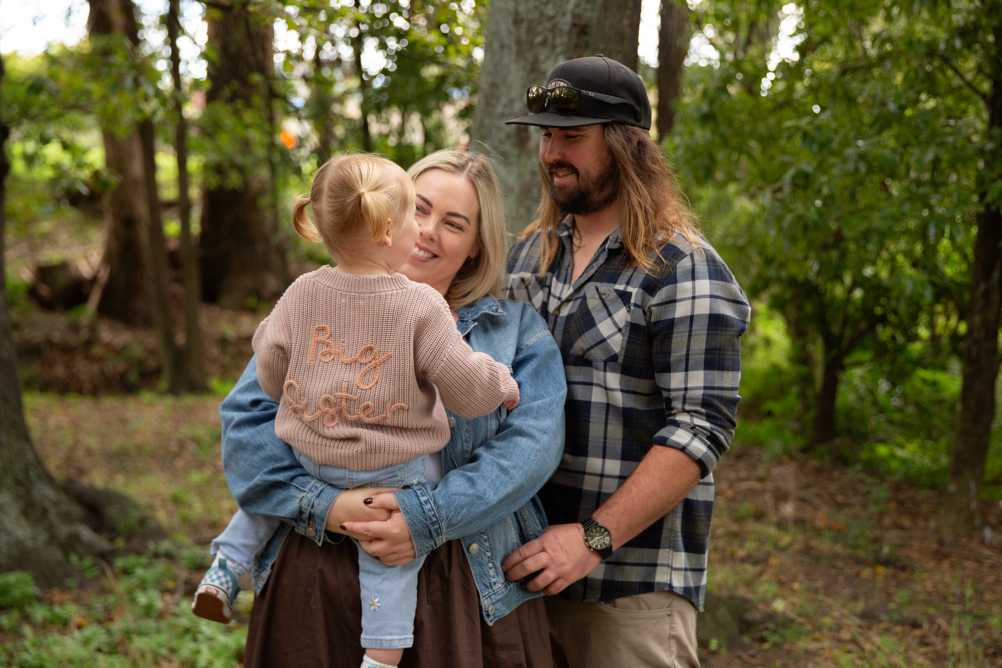 A family of three, including a young girl, standing outdoors in a wooded area, smiling and enjoying time together.