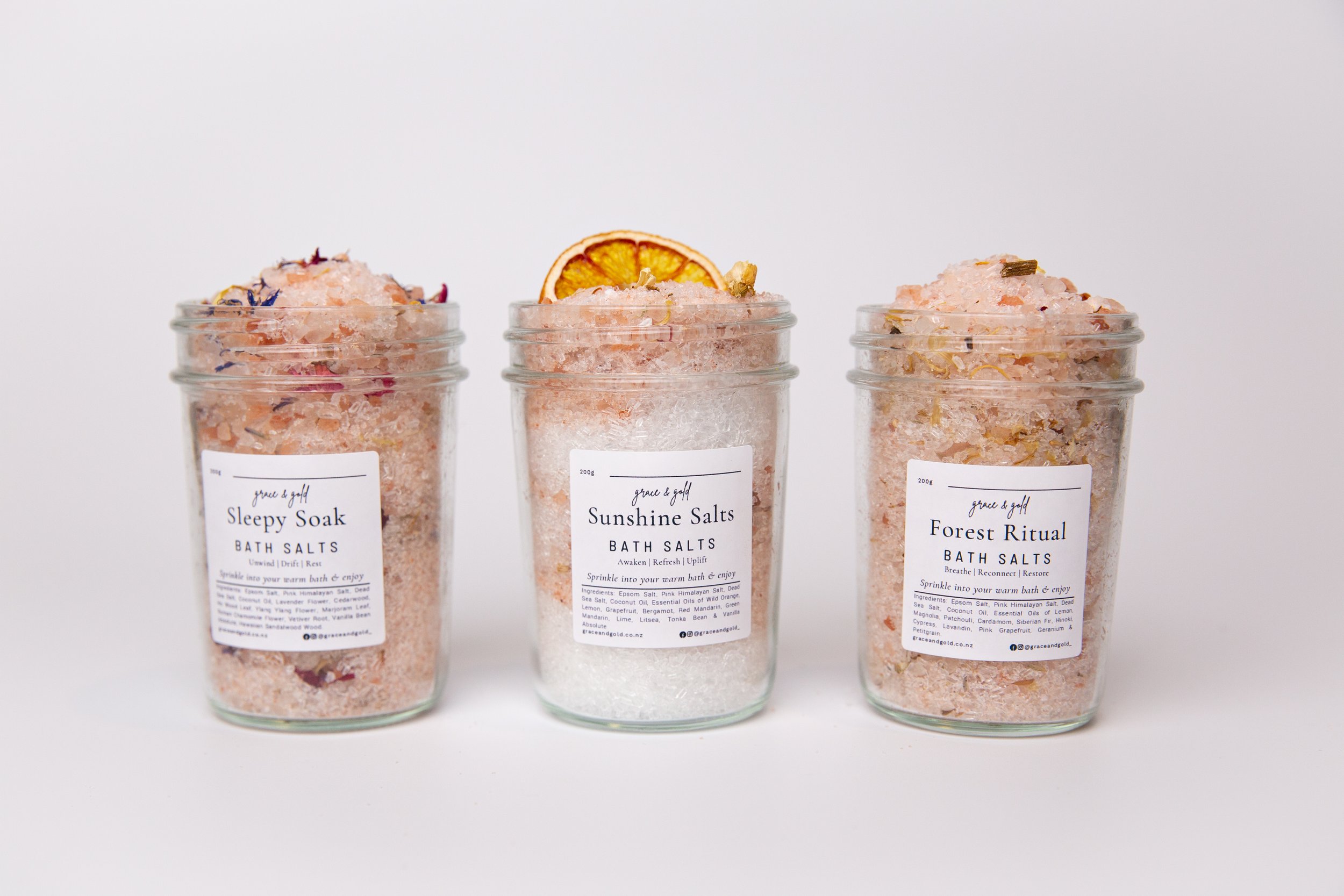 Three glass jars filled with bath salts, each labeled with different names: 'Sleepy Soak,' 'Sunshine Salts,' and 'Forest Ritual,' with the middle jar topped with a slice of dried orange.