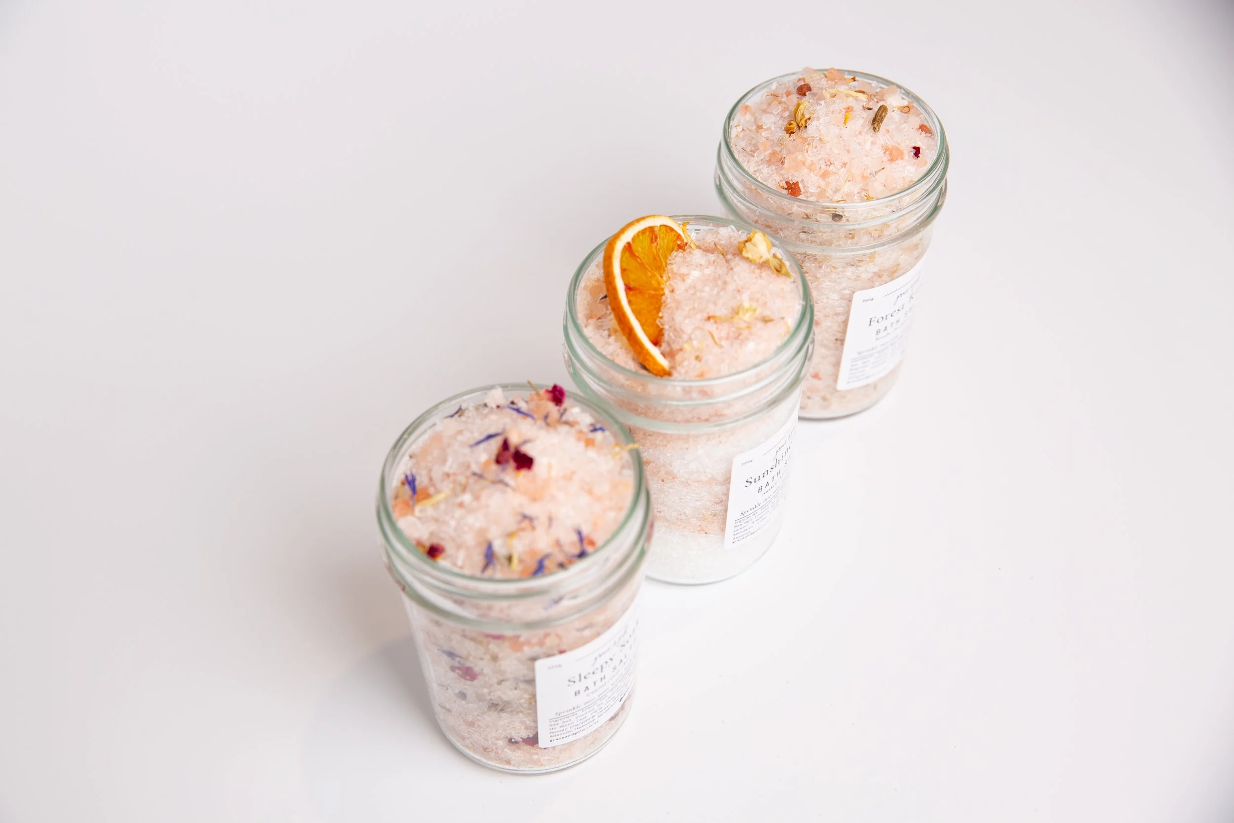 Four glass jars filled with pink bath salts topped with dried flower petals, a slice of dried orange on top of the salts, and a small paper label on each jar.