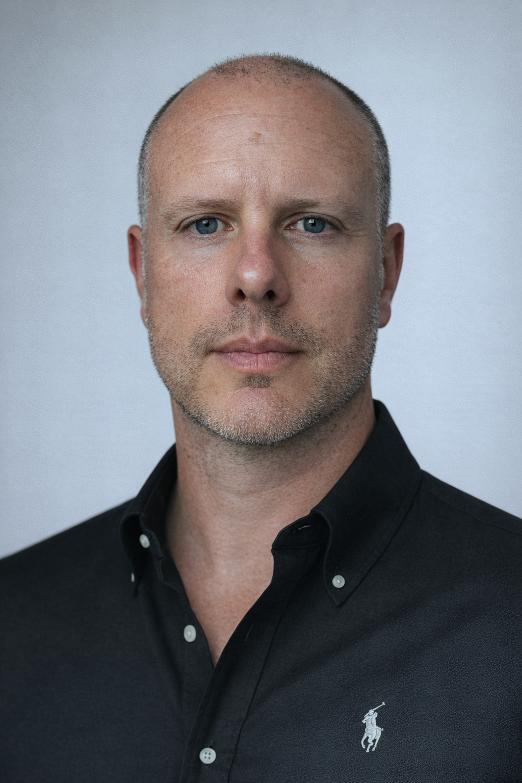 A man with short-cropped hair and blue eyes wearing a black collared shirt with a Polo Ralph Lauren logo.