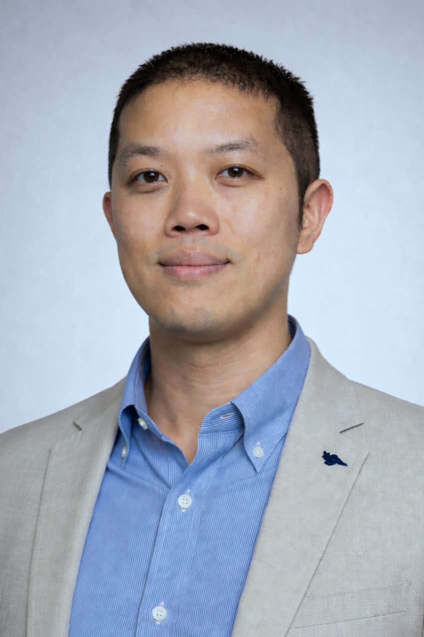 A professional headshot of an Asian man wearing a light beige suit jacket and a light blue button-down shirt, smiling slightly, with a plain light gray background.