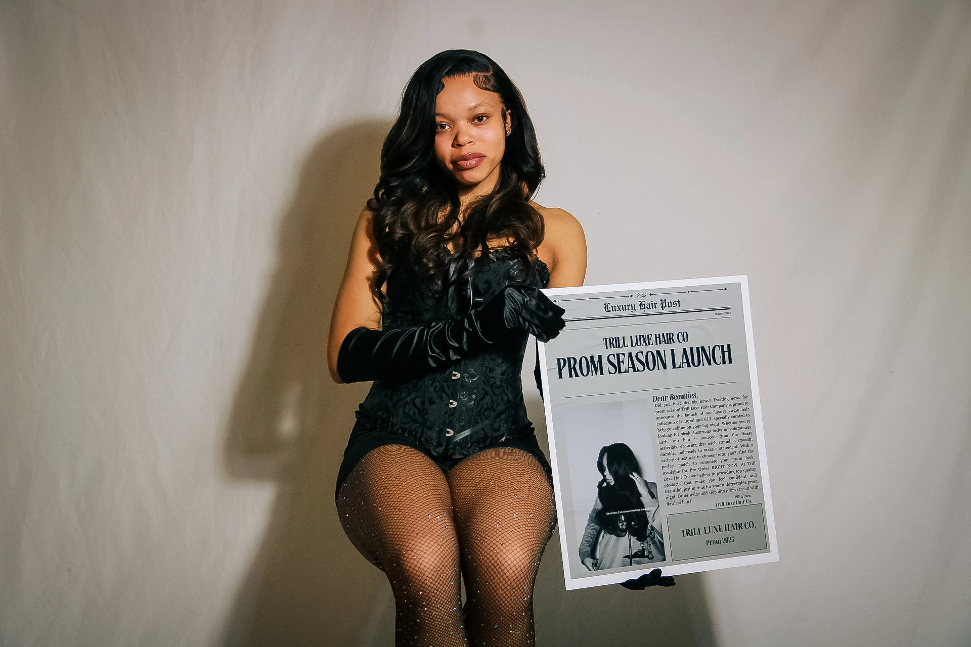 A woman dressed in black, wearing fishnet stockings with glitter, holding a newspaper that announces a prom season launch for Trill Luxe Hair Co. She is standing against a plain wall and looking at the camera.
