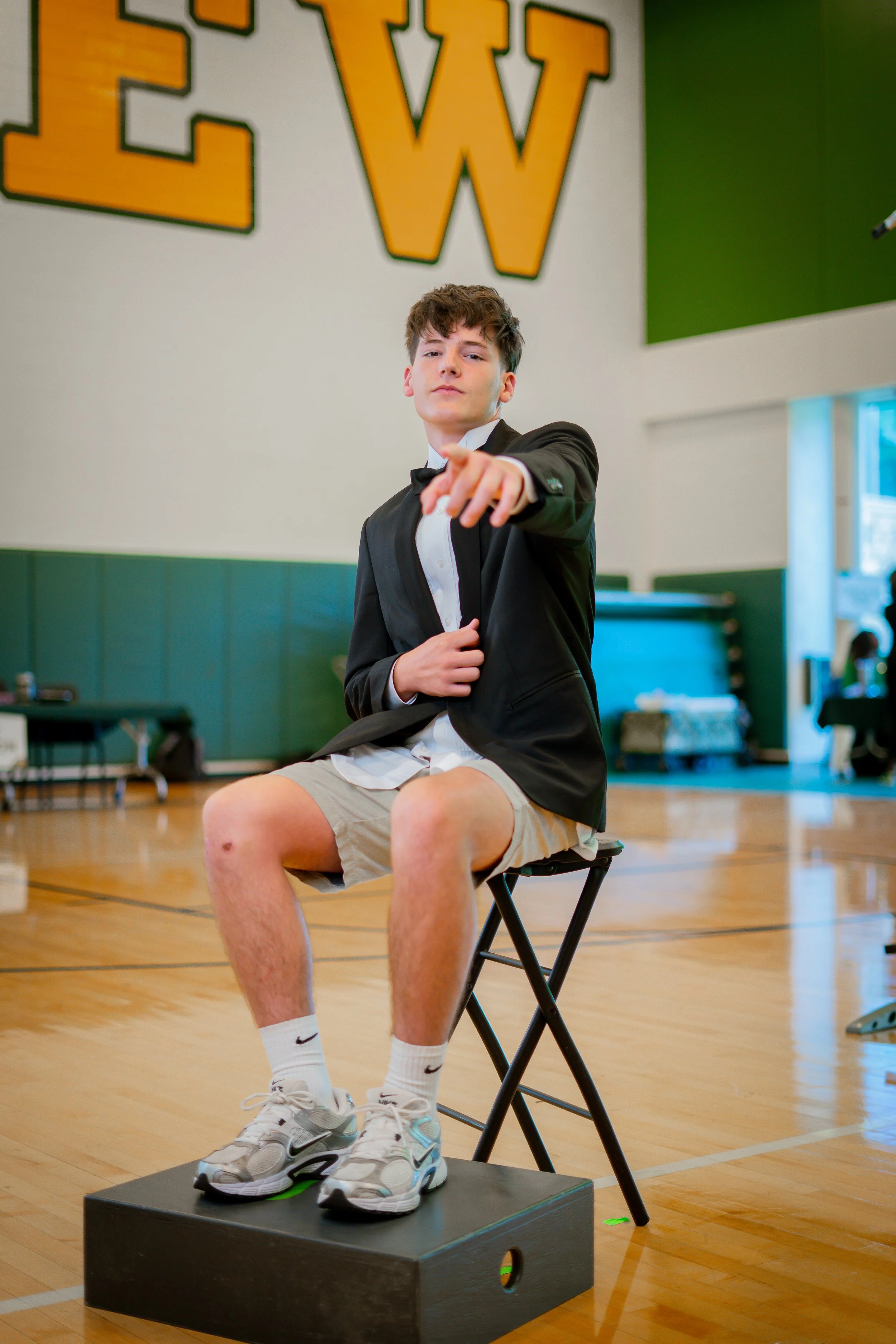 A young man in a tuxedo jacket, white shirt, and shorts sitting on a chair on a small platform in a gymnasium, pointing towards the camera with a confident expression.