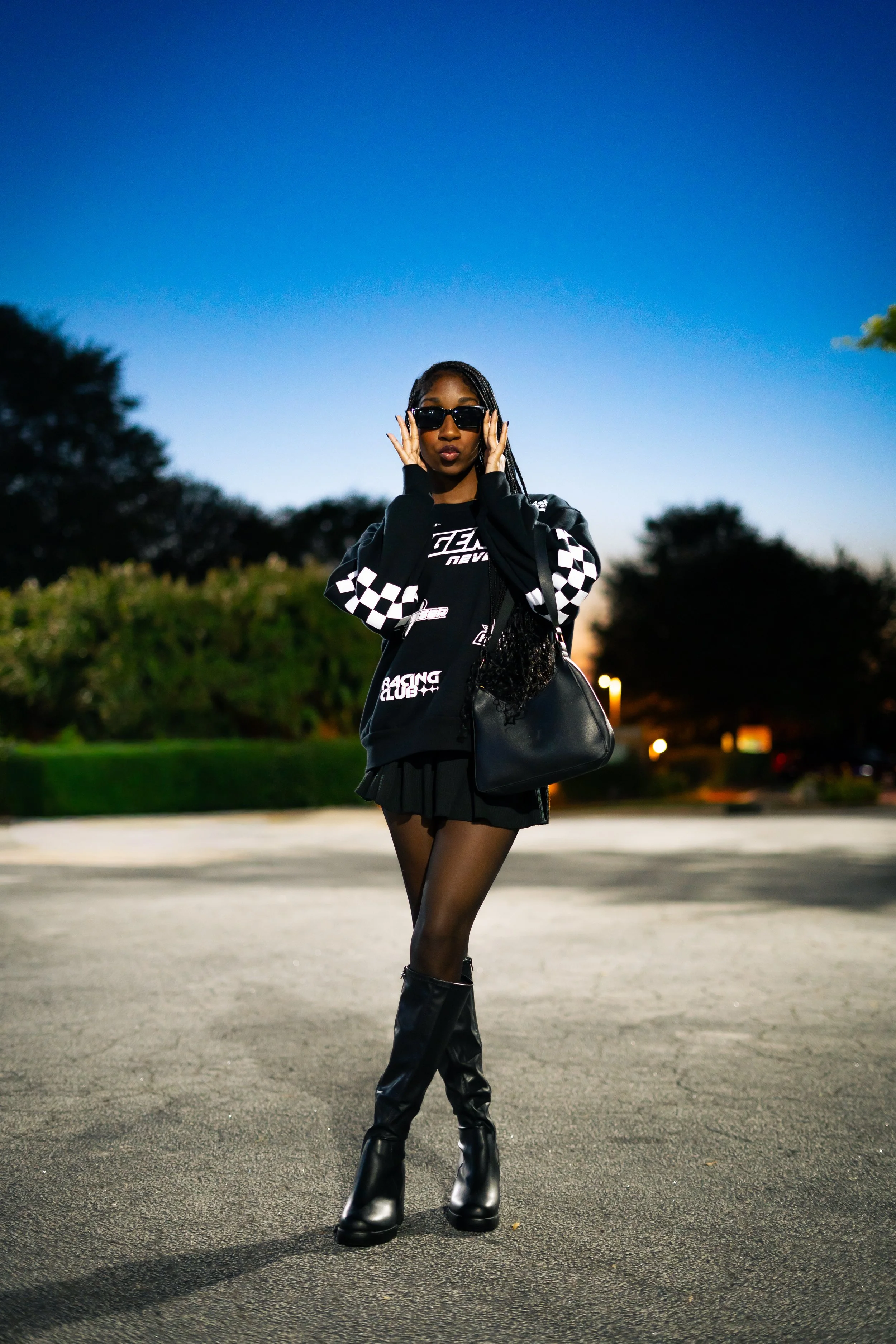 A fashionable young woman with braided hair, wearing sunglasses, a black racing-themed hoodie, a short skirt, sheer black tights, and knee-high black boots, stands outdoors on an empty parking lot at dusk, posing with her hands near her face.