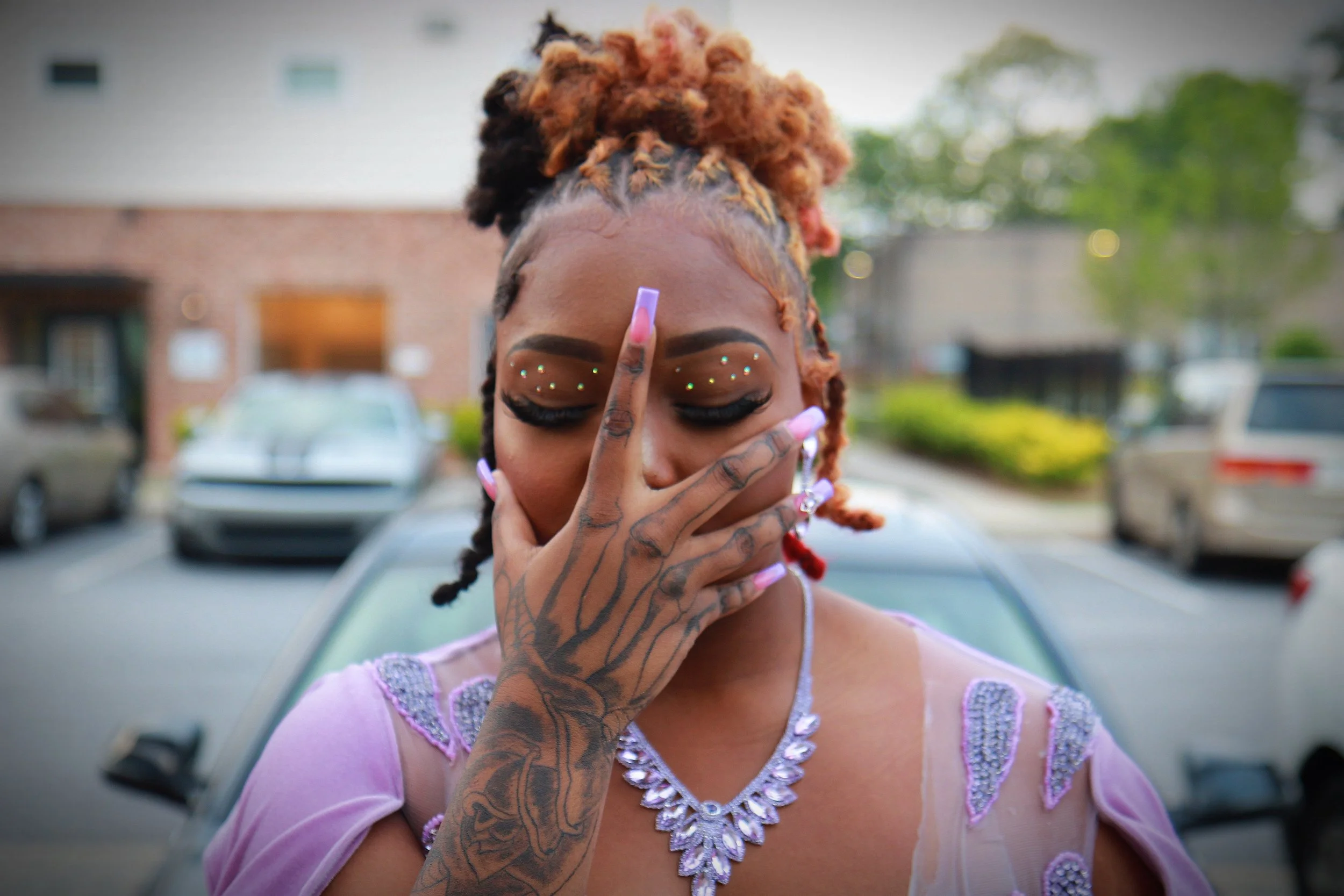 A woman with dyed orange and black dreadlocks, wearing makeup with rhinestones, covering her face with her hand and eyes closed in a parking lot.