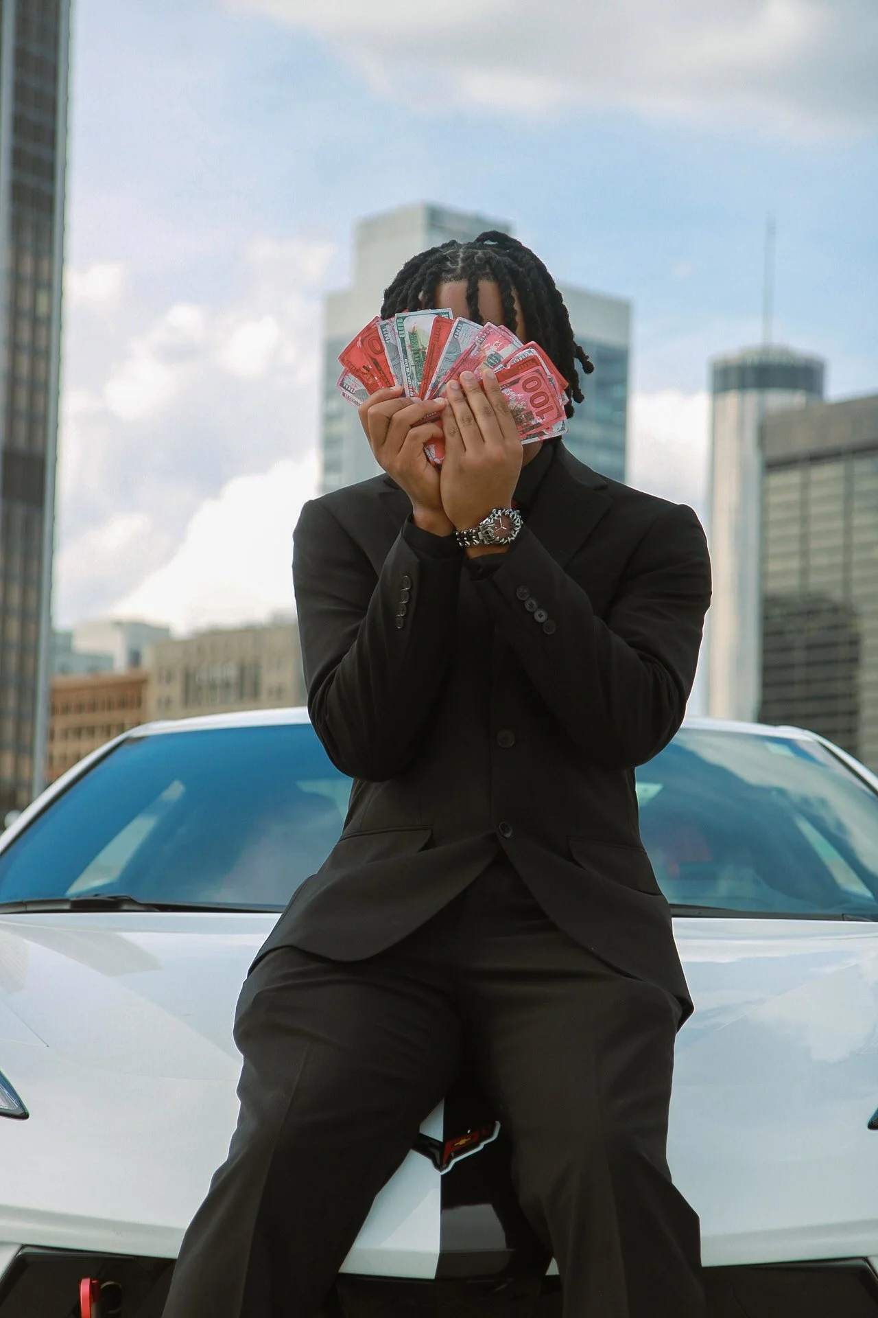 A person in a black suit holding a fan of red and green currency notes in front of their face, leaning against a white sports car in an urban setting with tall buildings in the background.