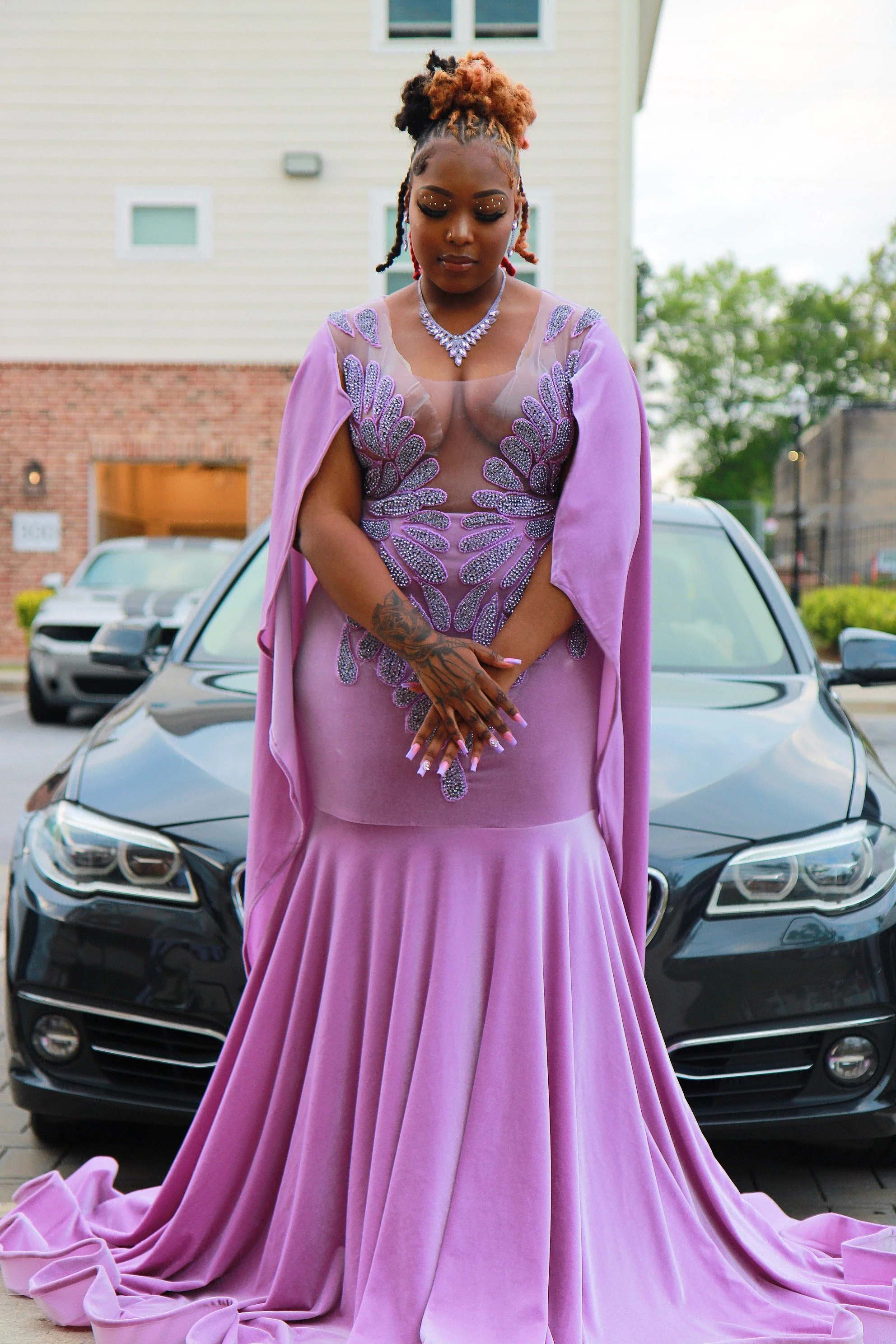 A woman standing in front of a black car, wearing a purple evening gown with bead and sequin embellishments. She has braided hair styled into an updo and is accessorized with jewelry. Her hands are clasped in front of her.