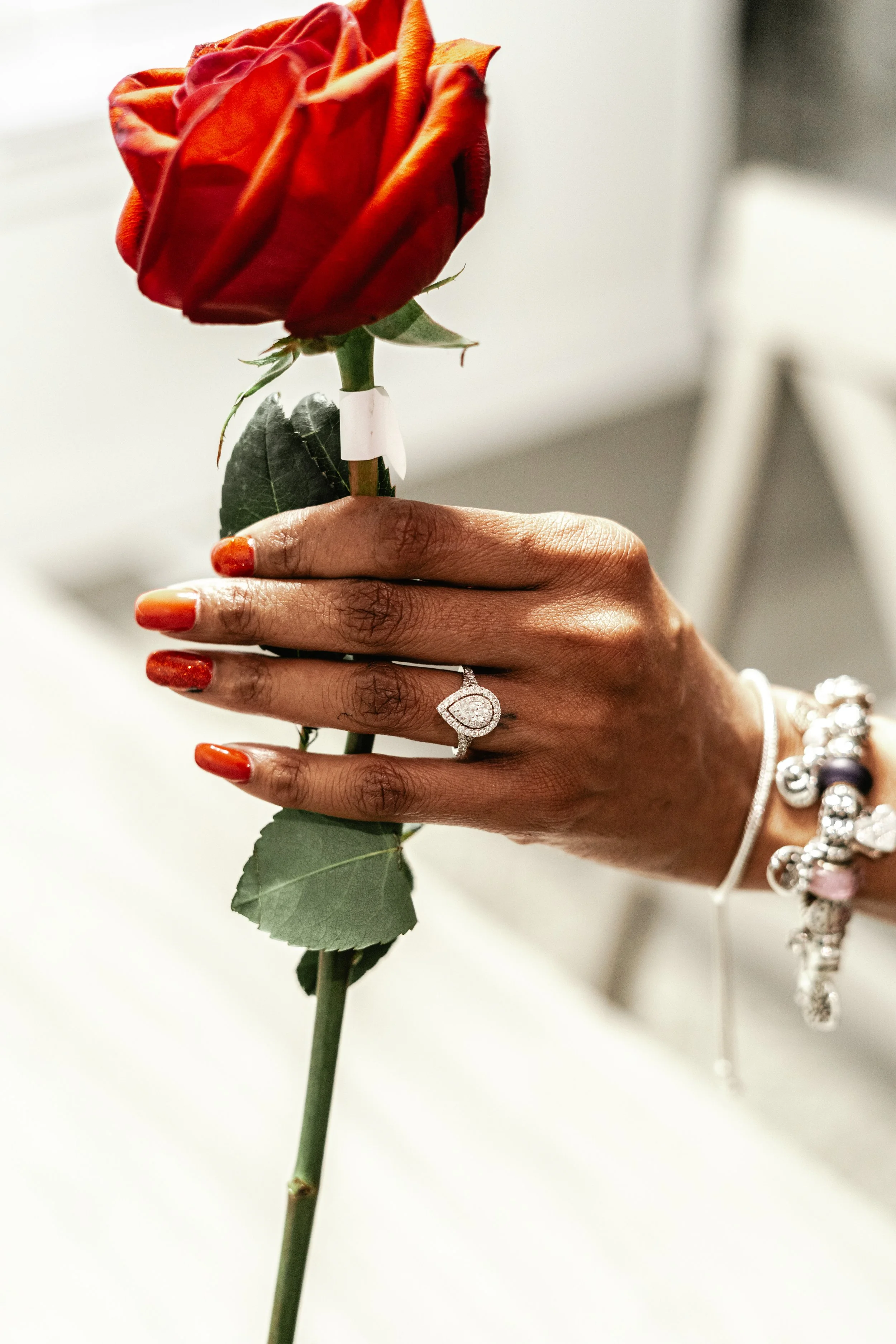 A hand with orange painted nails holding a red rose with a diamond ring on the ring finger, wearing bracelets.