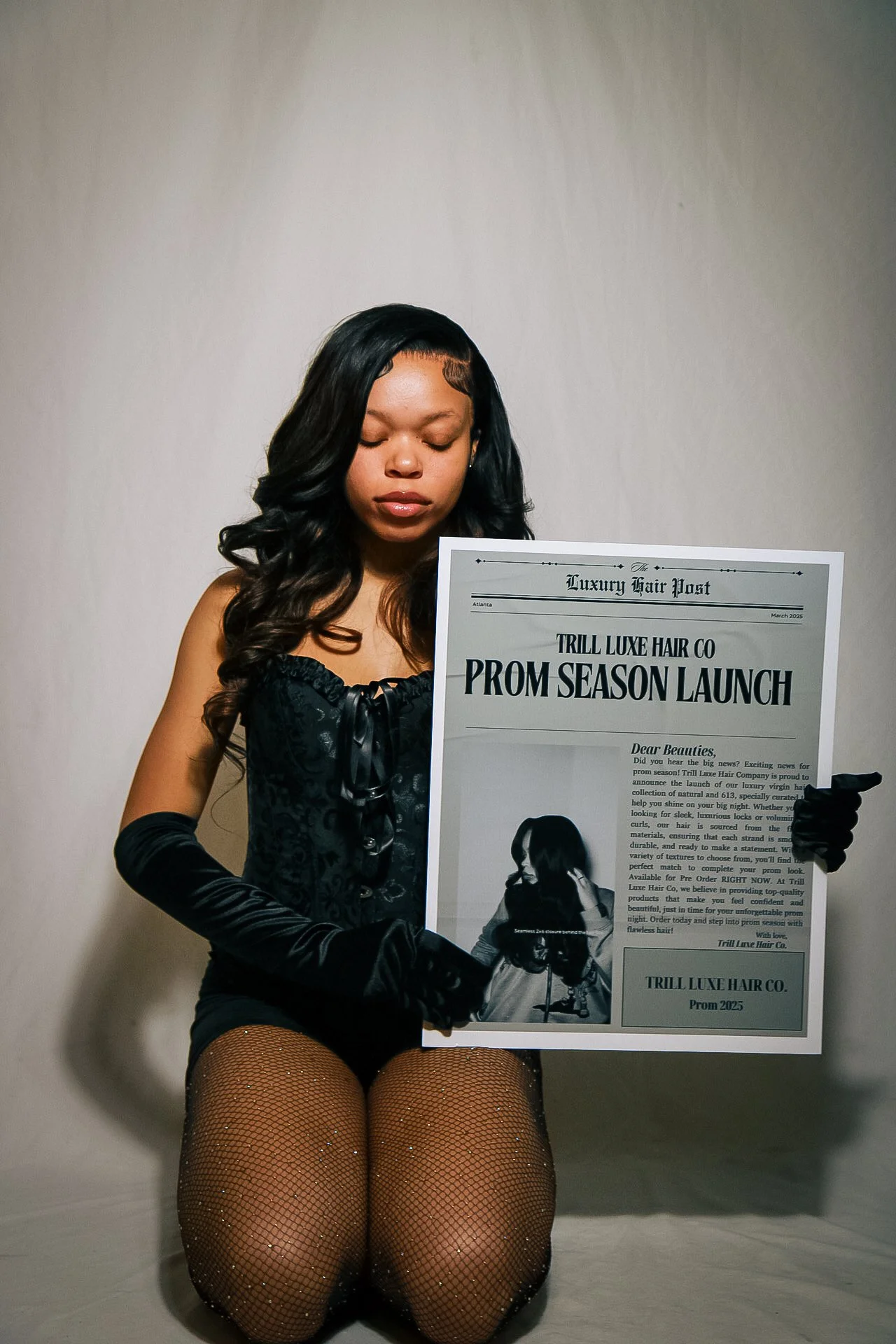 A woman dressed in black lace outfit with long gloves, fishnet stockings, and styled hair kneels on the floor holding a large promotional poster for a prom season hair launch event.