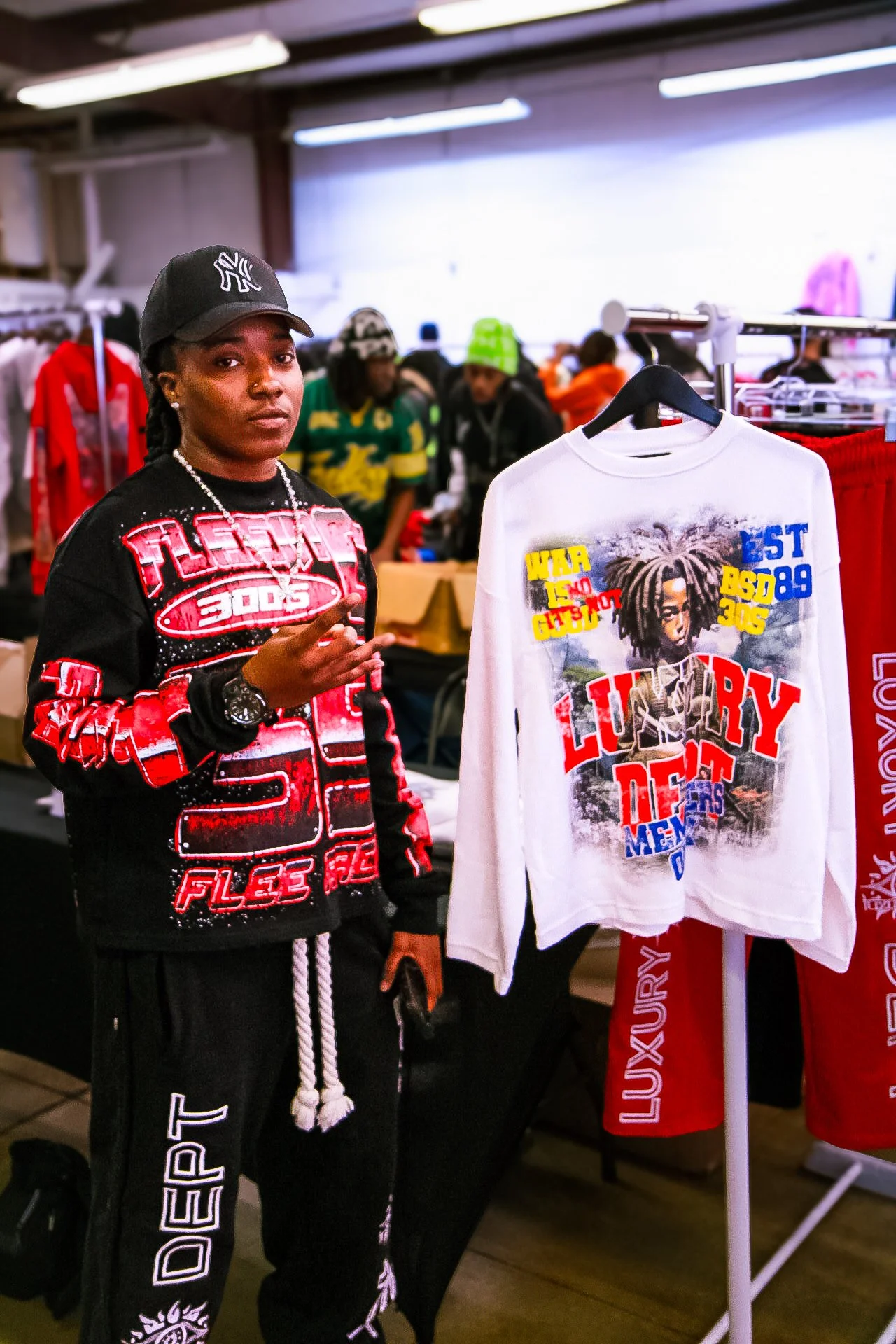 A young woman with dark hair and wearing a black cap, black and red sweatshirt, and black pants, stands next to a white sweatshirt with colorful graphics and text on a clothing rack at an indoor market or store.