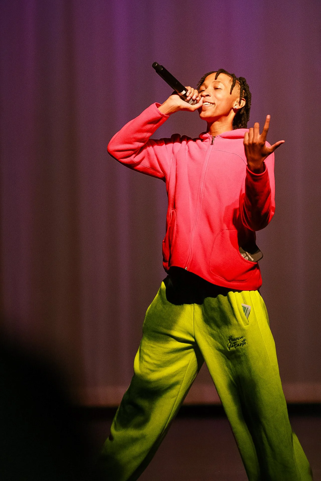 A young person singing into a microphone on stage, wearing a bright pink zip-up jacket and yellow-green pants.