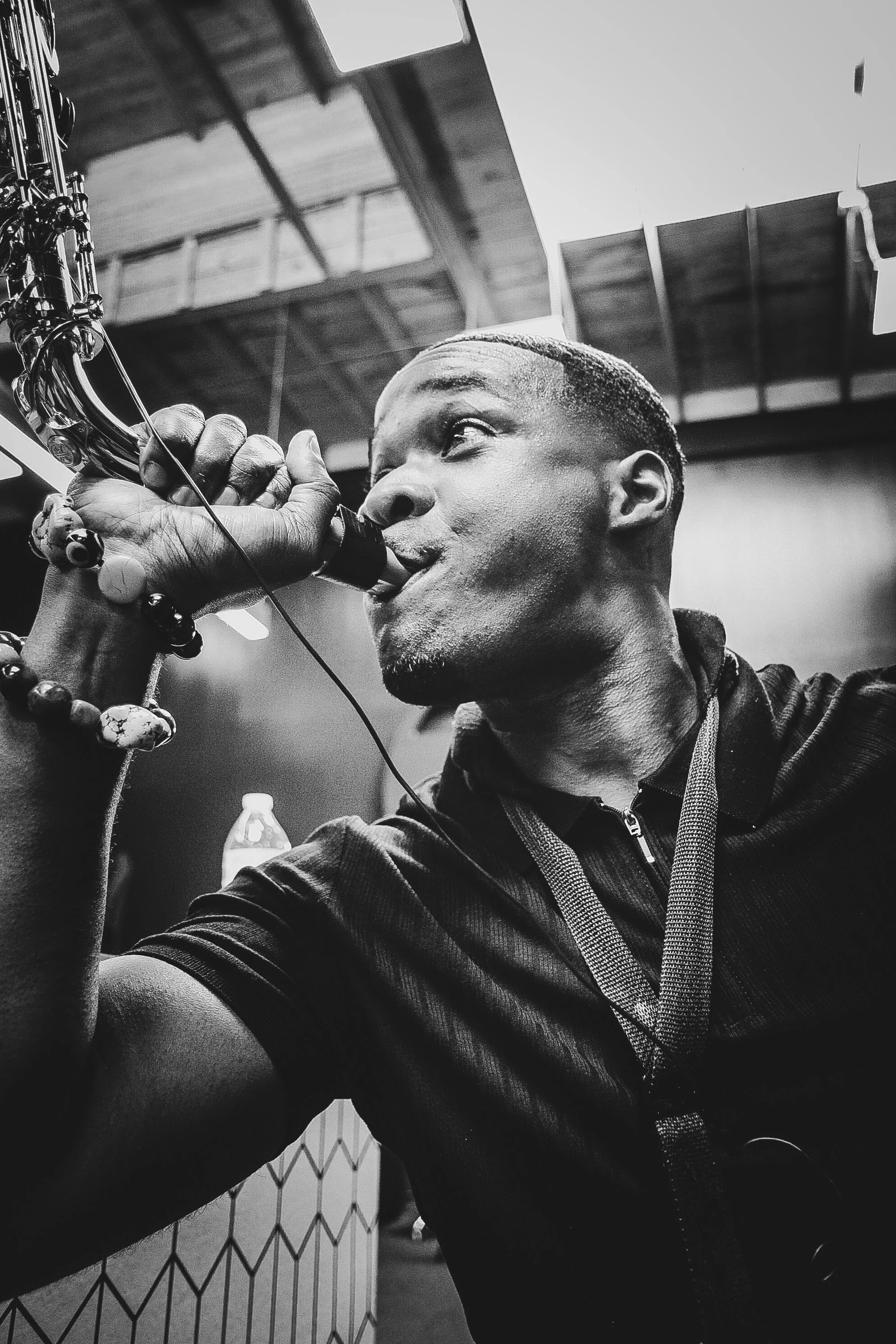 A black and white photo of a man playing a saxophone, taken from a low angle, with a ceiling and some background objects visible.