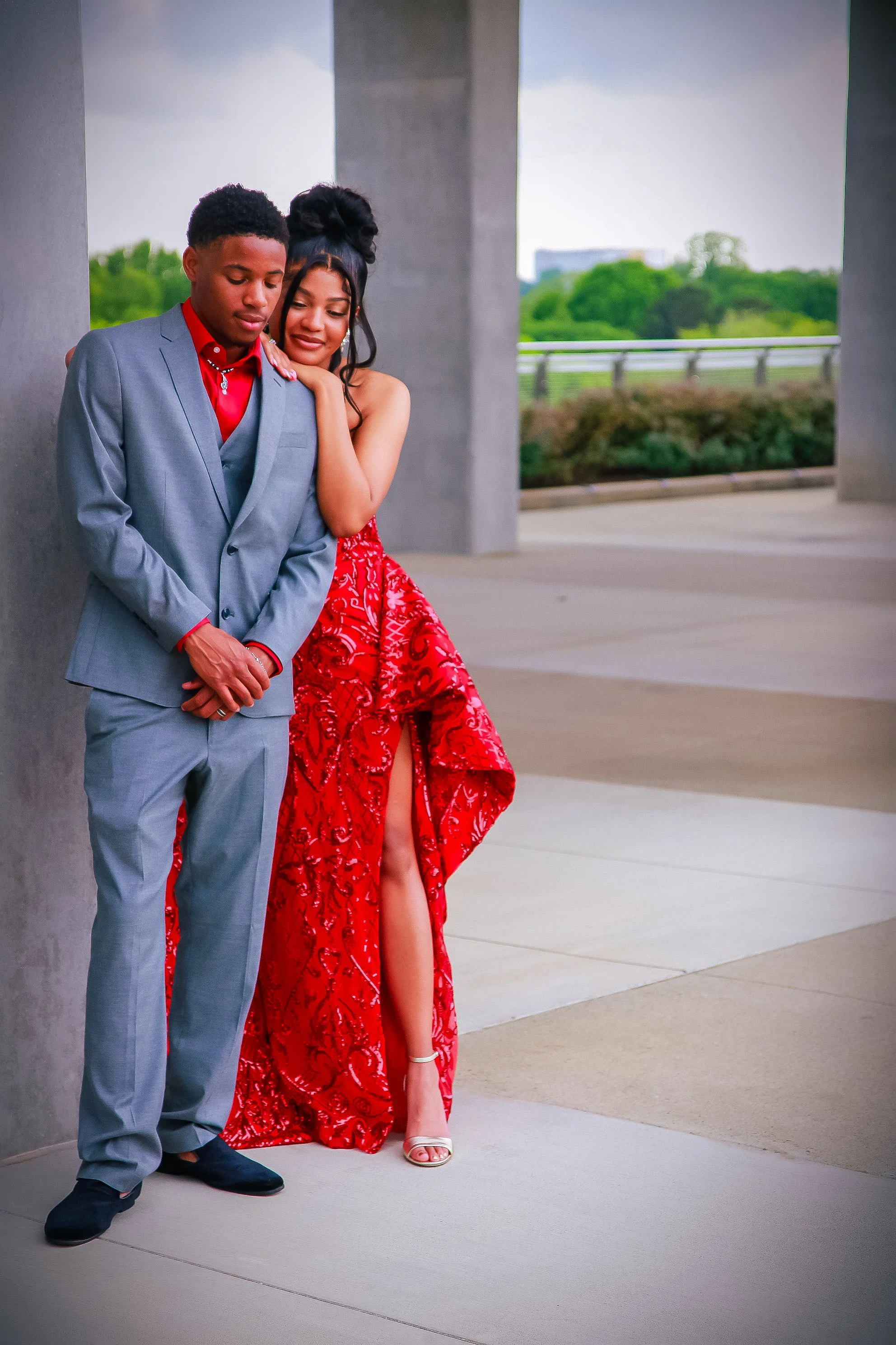 A young man in a gray suit and red shirt standing with a young woman in a red dress with a high slit, both leaning against a concrete pillar outdoors. She is gently touching his shoulder while he looks down, and she is smiling softly.