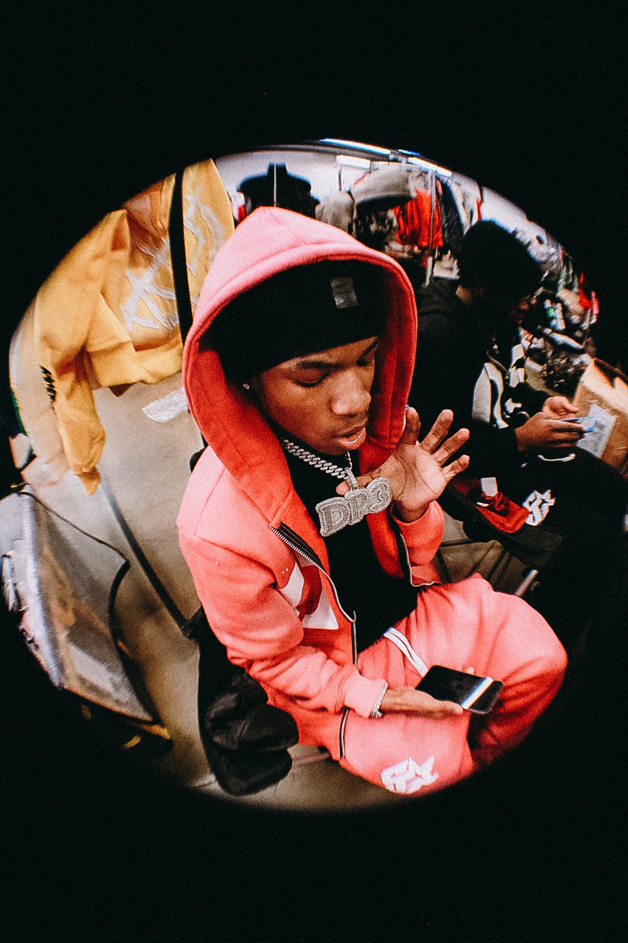 A young man wearing a pink hoodie, black beanie, and large chain with a pendant, sitting on a chair, holding a phone, in an indoor setting with people and clothing in the background.