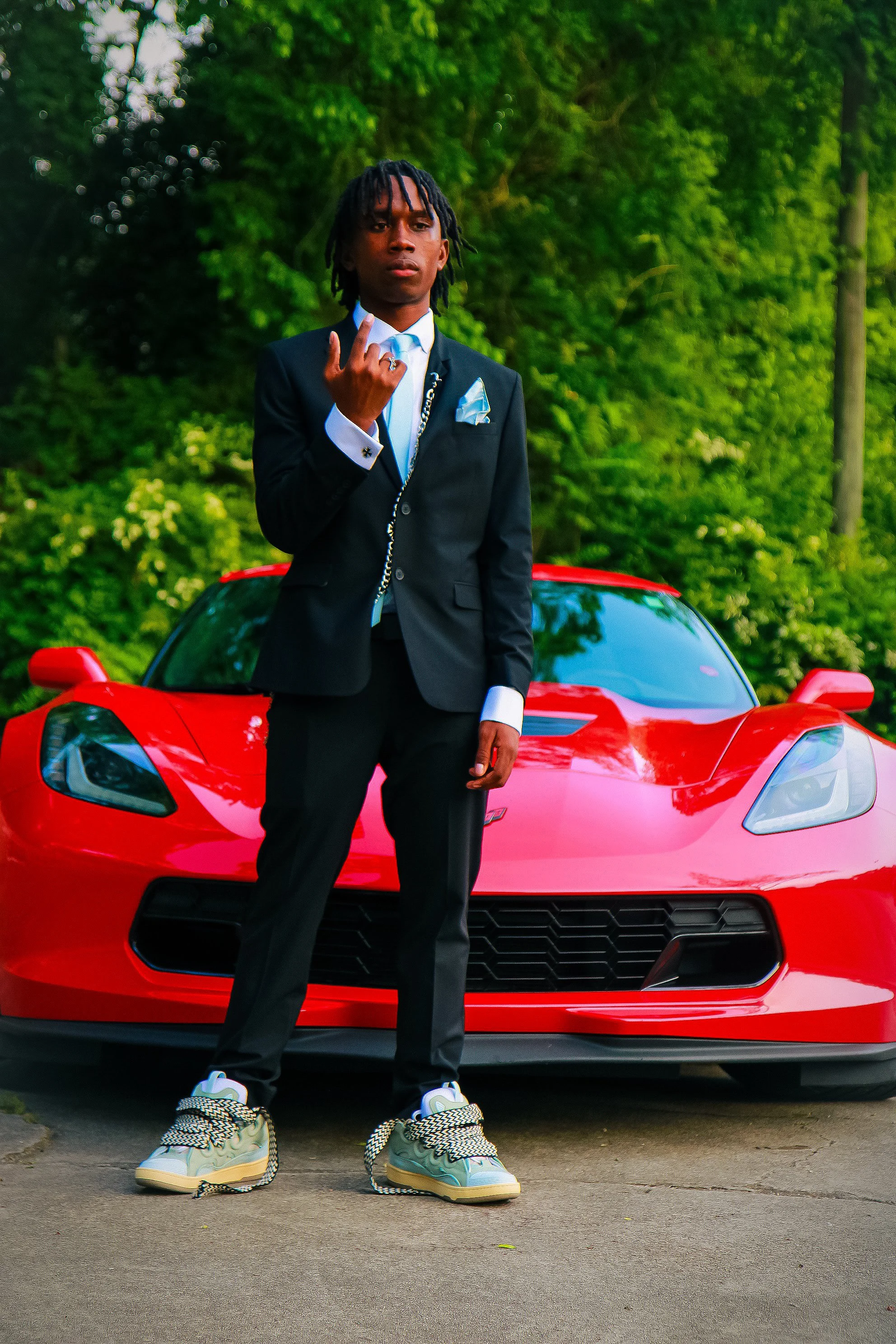 A young man in a black suit with a light blue tie and pocket square stands in front of a red sports car, making a hand gesture, outdoors with green trees in the background.