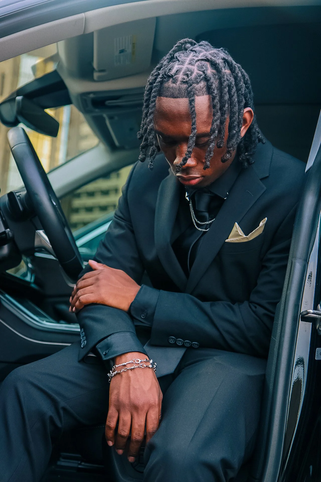 A man dressed in a black suit, sitting inside a vehicle, with his head bowed and eyes closed, wearing jewelry and a gold pocket square.