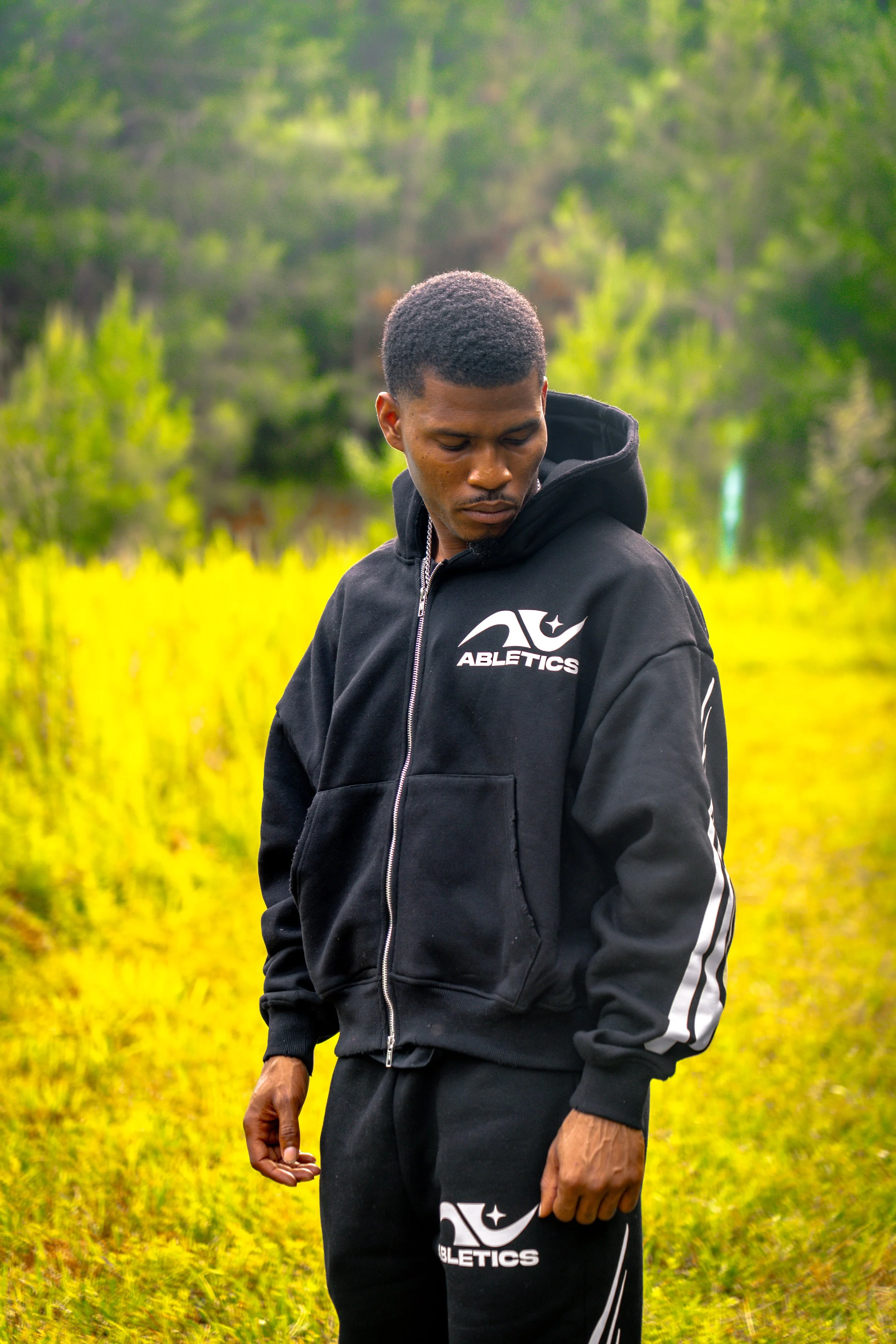 A young man in a black athletic hoodie and pants standing outdoors in a grassy field with trees in the background.