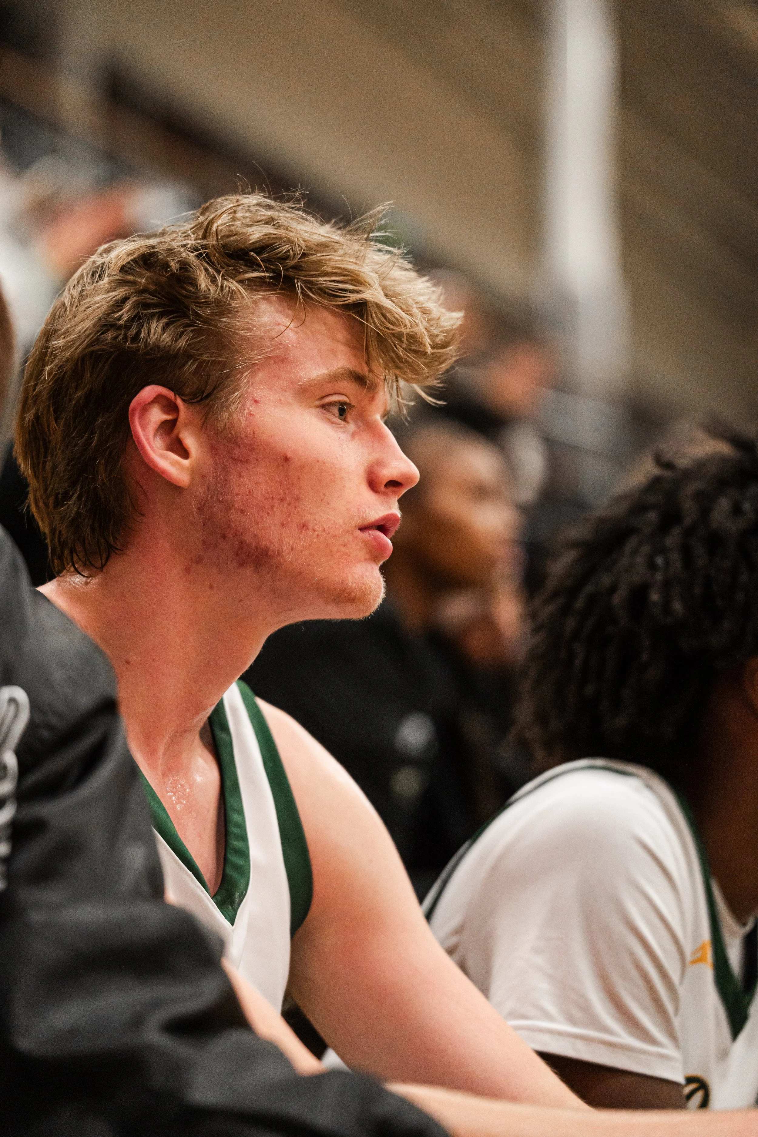 A young male basketball player with reddish hair and acne, wearing a white jersey with green trim, sitting on the sidelines during a game.