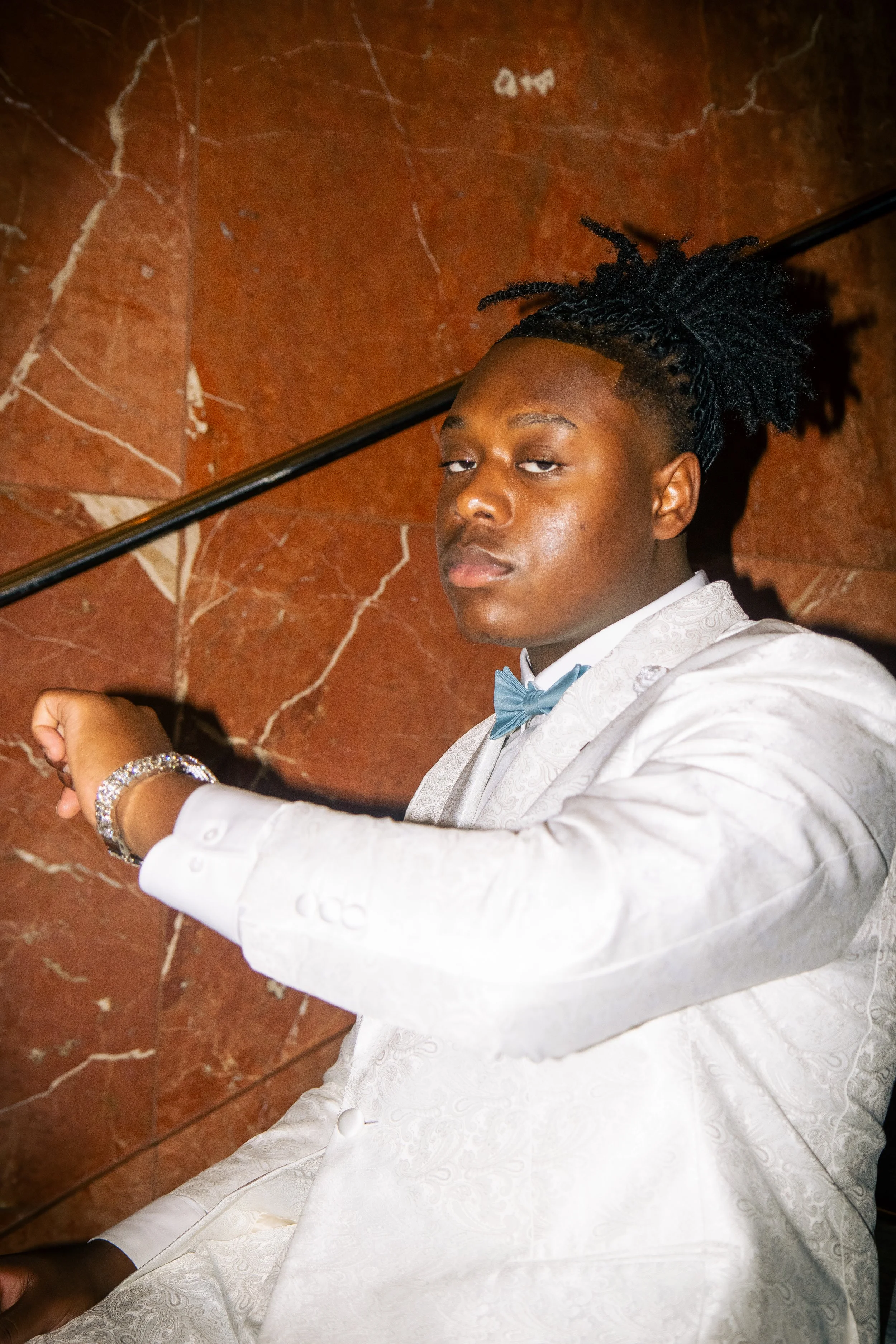 Young man in a white tuxedo with a blue bow tie sitting against a marble wall, wearing a bracelet, with styled dreadlocks.