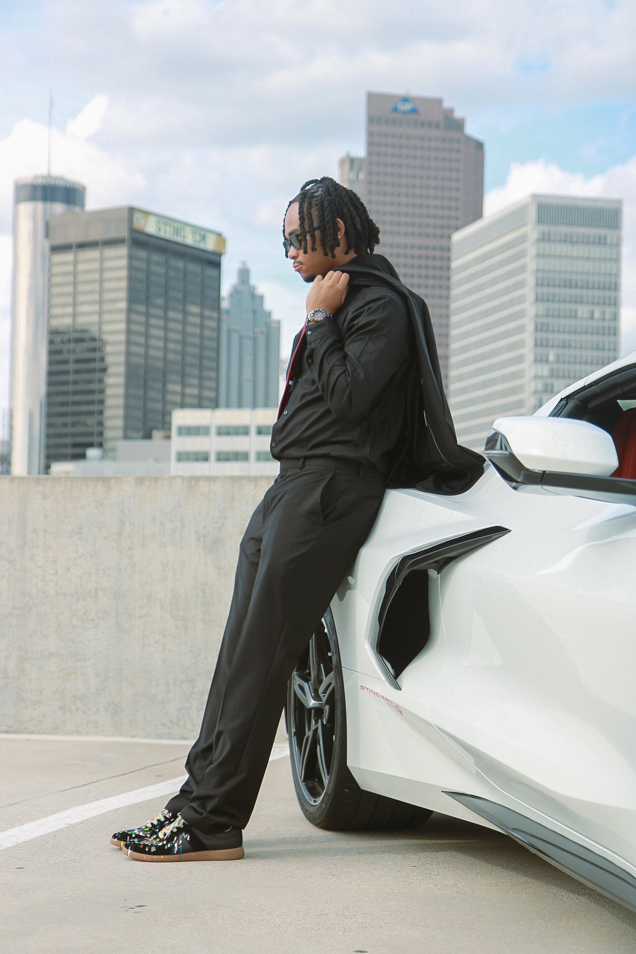 A man with dreadlocks leaning against a white sports car on a rooftop parking lot with a city skyline in the background.