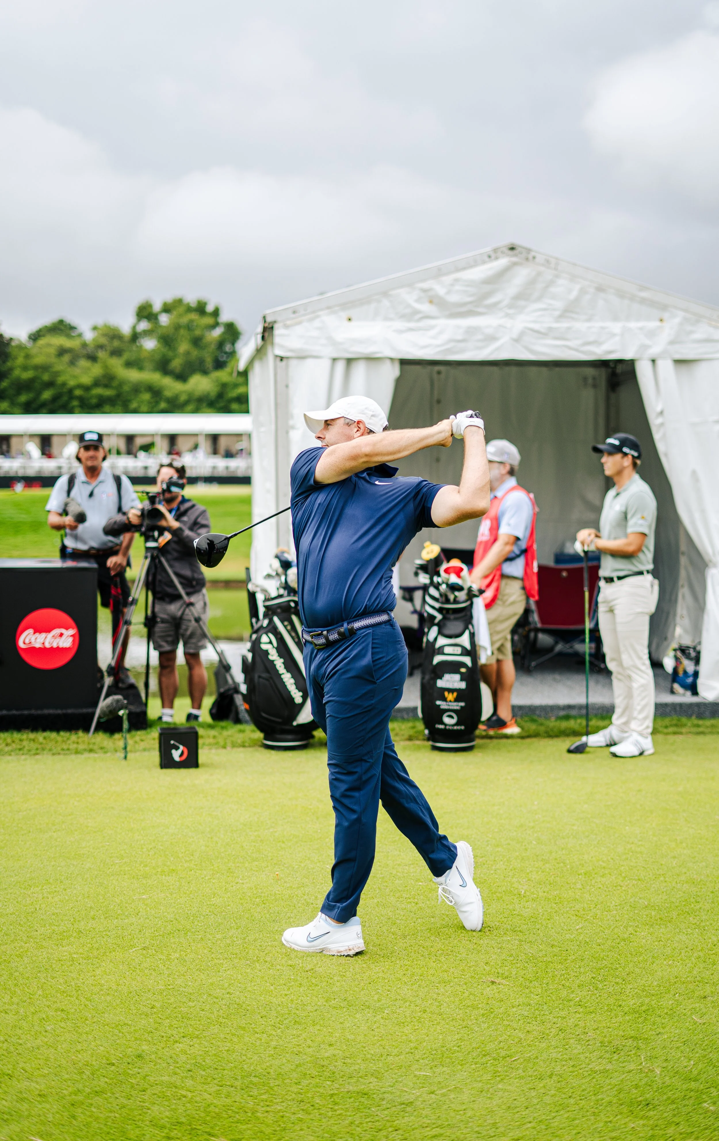 A golfer practicing his swing on a golf course, with a white tent and other golfers and camera crew in the background.