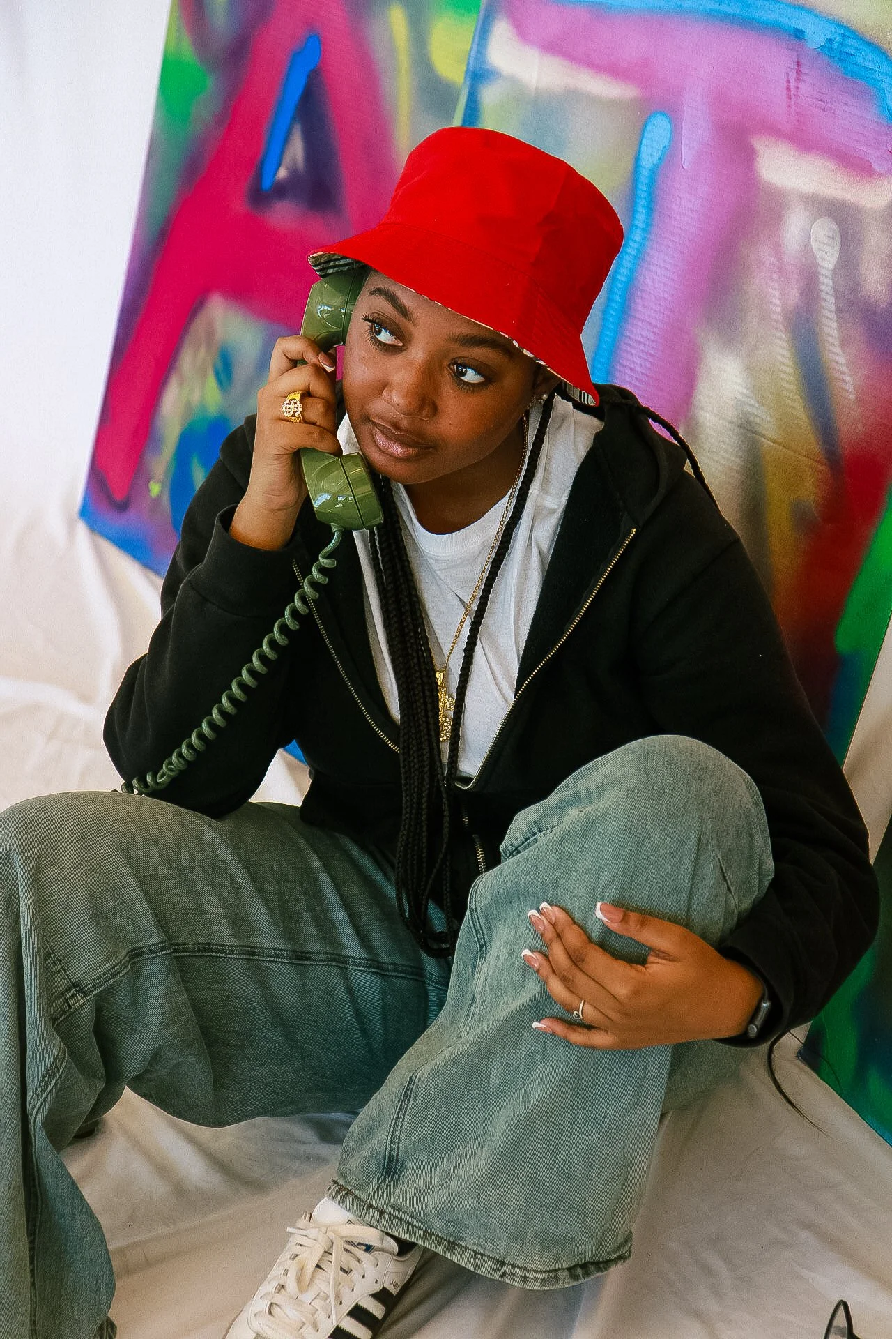 Young woman with braids, wearing a red hat, black hoodie, gray jeans, and white sneakers, sitting on the floor, talking on a green vintage telephone, with colorful abstract paintings in the background.