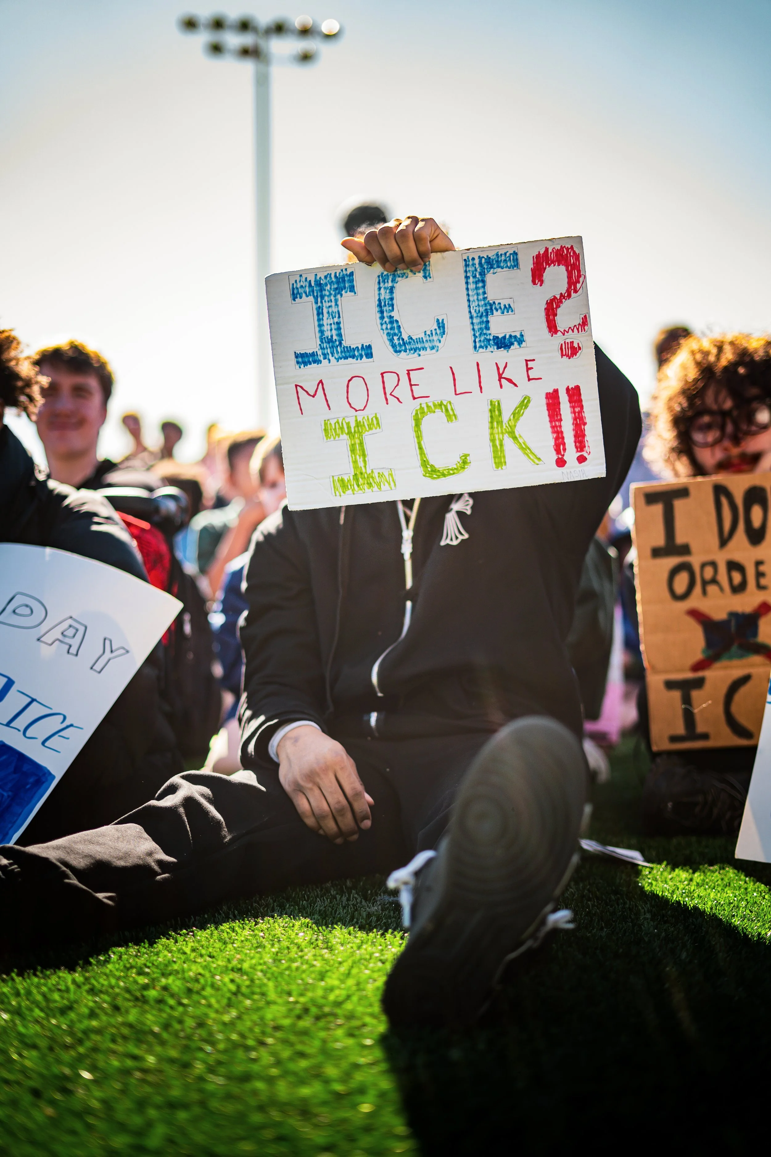 Protesters gathered outdoors, with one person sitting on the grass holding a colorful hand-made sign that says, 'ICE? More Like TICK!!' in large, colorful letters. There are other protest signs visible, and the crowd is illuminated by bright sunlight