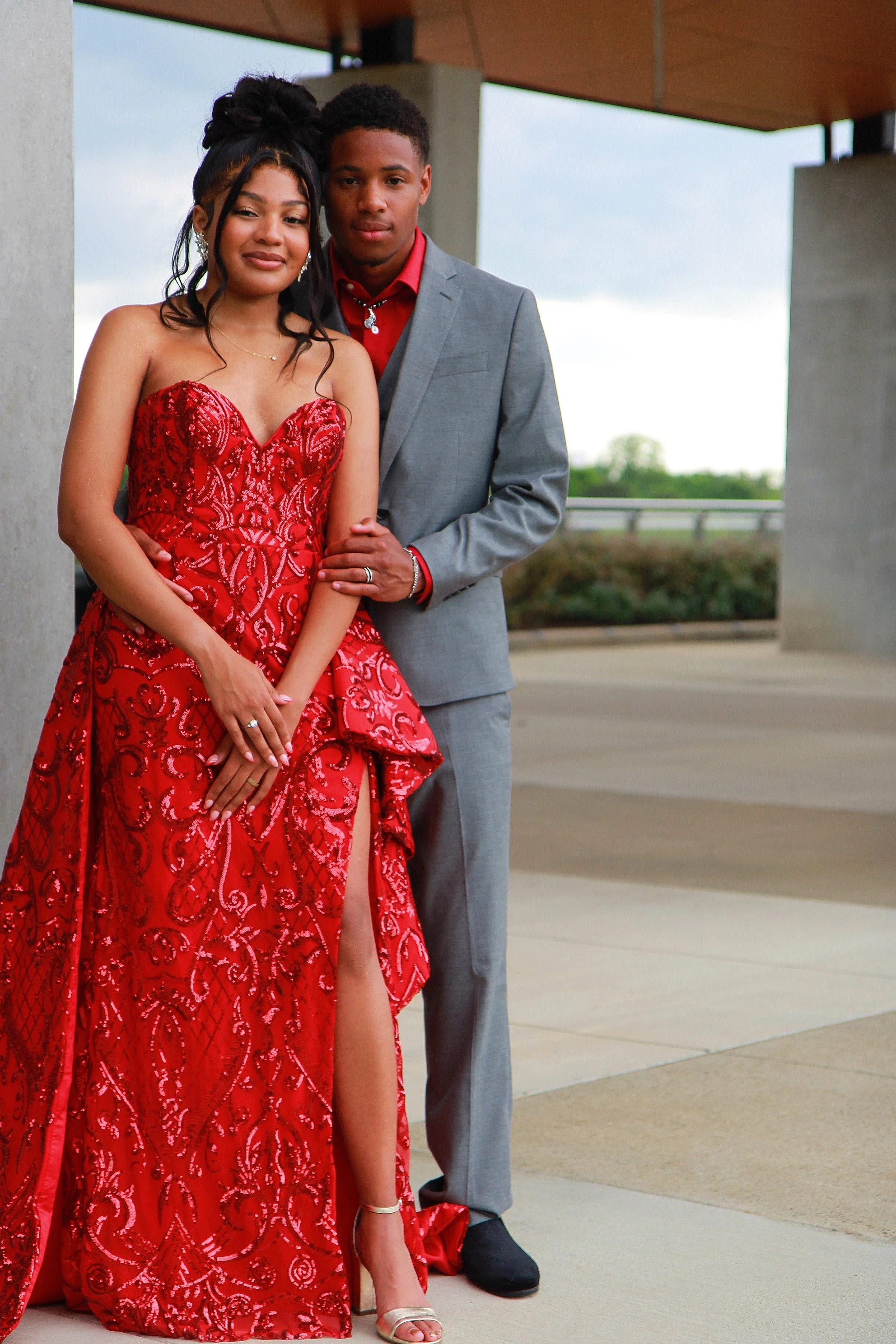 A young couple dressed in formal attire standing outdoors, with the woman wearing a red evening gown and the man in a gray suit.