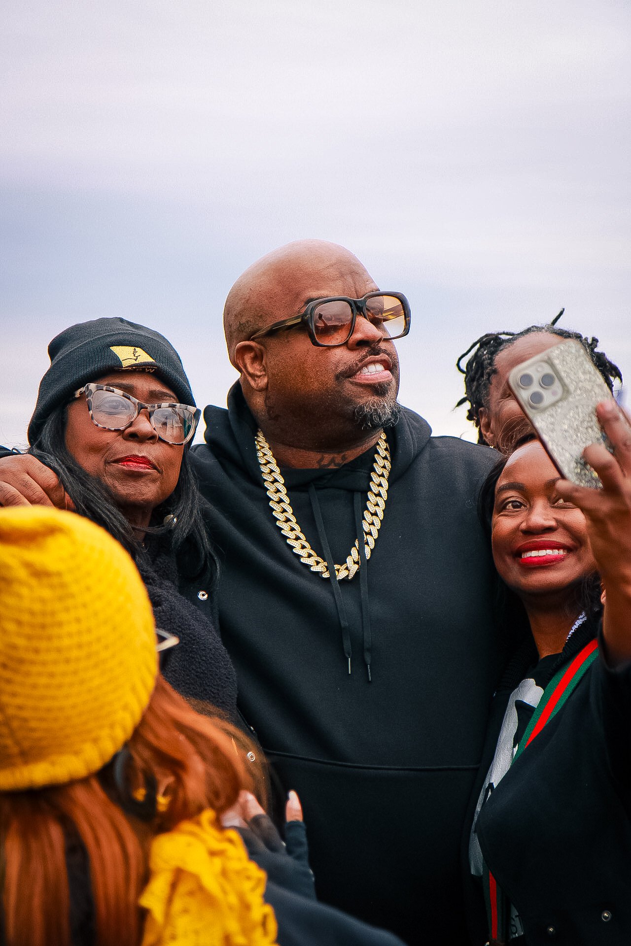 A group of four people taking a selfie outdoors, dressed warmly, with a cloudy sky in the background. The central man is wearing large glasses, a black hoodie, and a gold chain, while the women around him are smiling and wearing glasses and warm clot