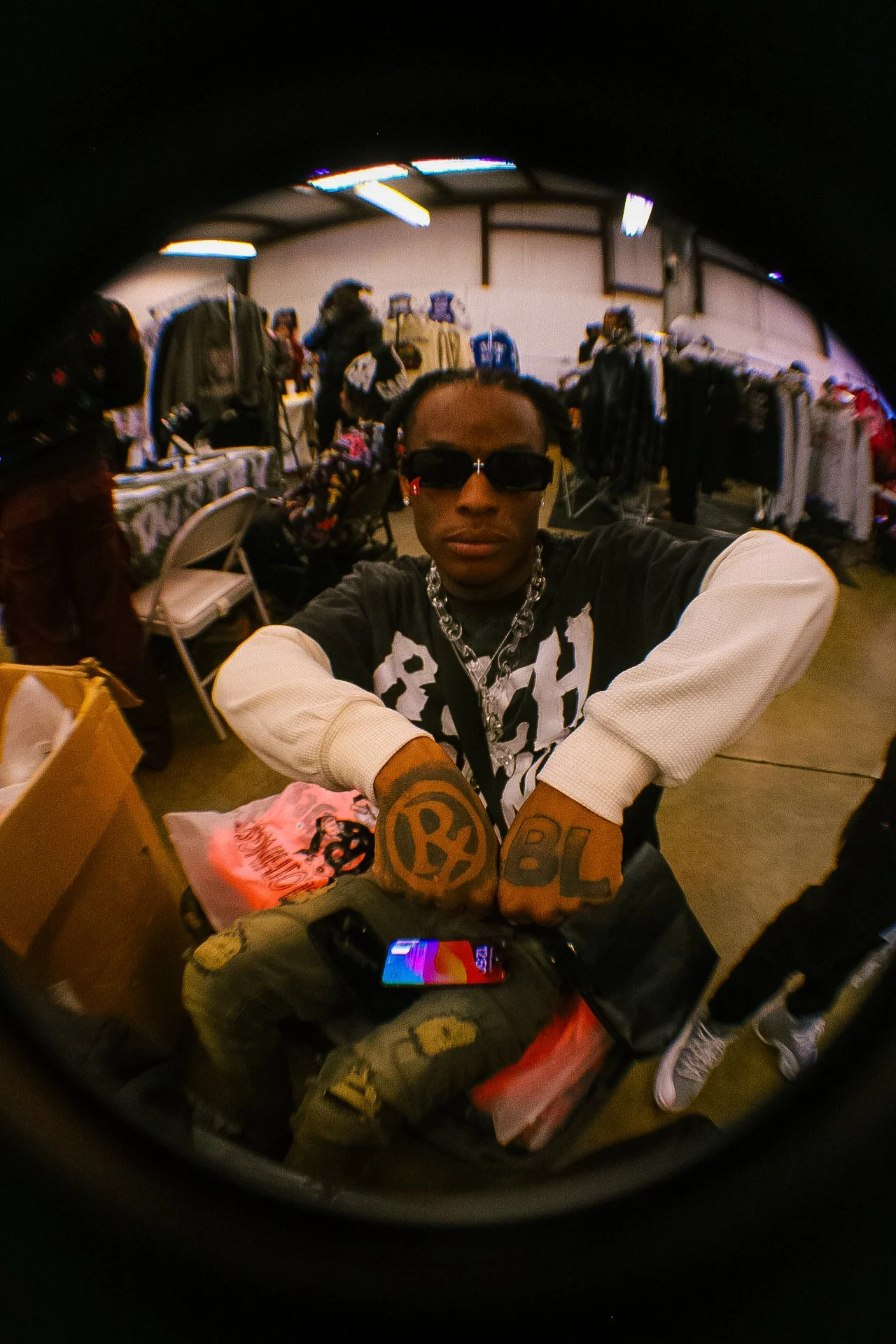 Person wearing sunglasses and a chain necklace sitting at a table in a clothing store, taking a selfie with a fisheye lens. The store has clothing racks and other shoppers in the background.