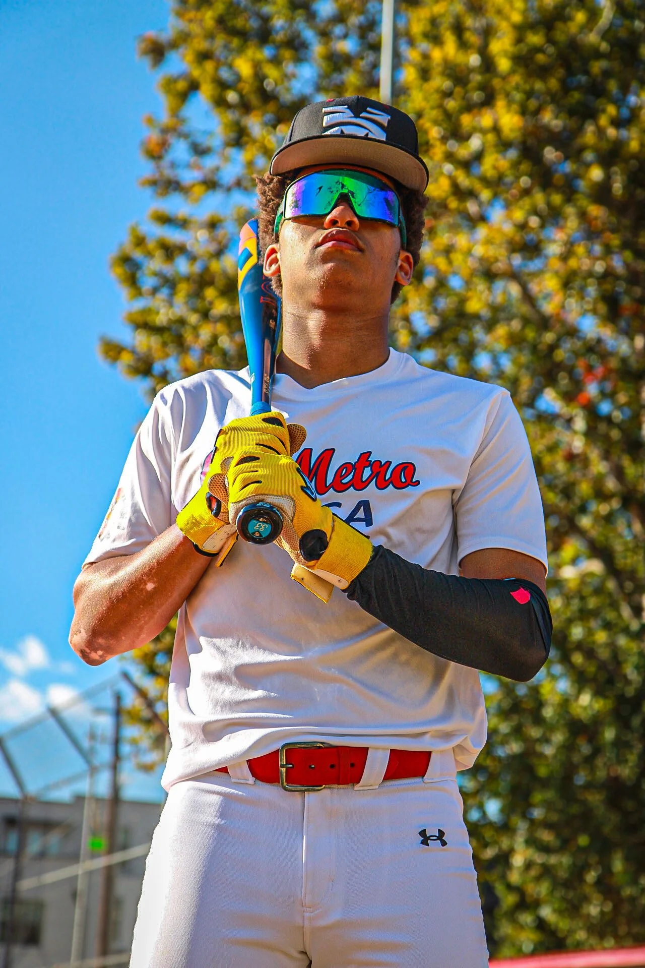 Young male baseball player wearing sunglasses, cap, and white uniform holding a bat on his shoulder, standing outdoors with trees and blue sky in the background.