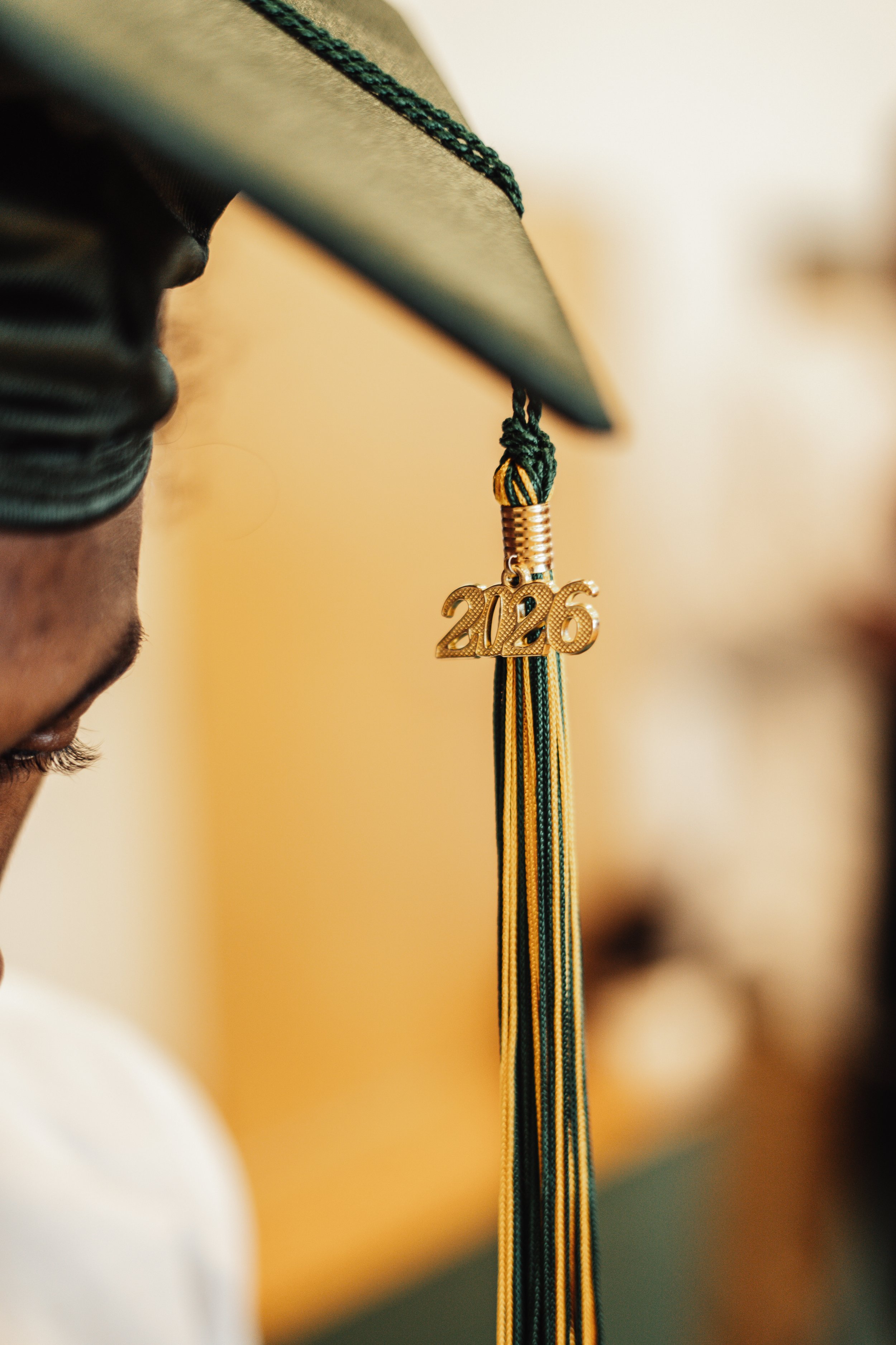 Close-up of a graduation cap with a tassel that has a gold "2026" charm, beads, and ribbons hanging from it, worn by a person with dark skin, and part of their face is visible