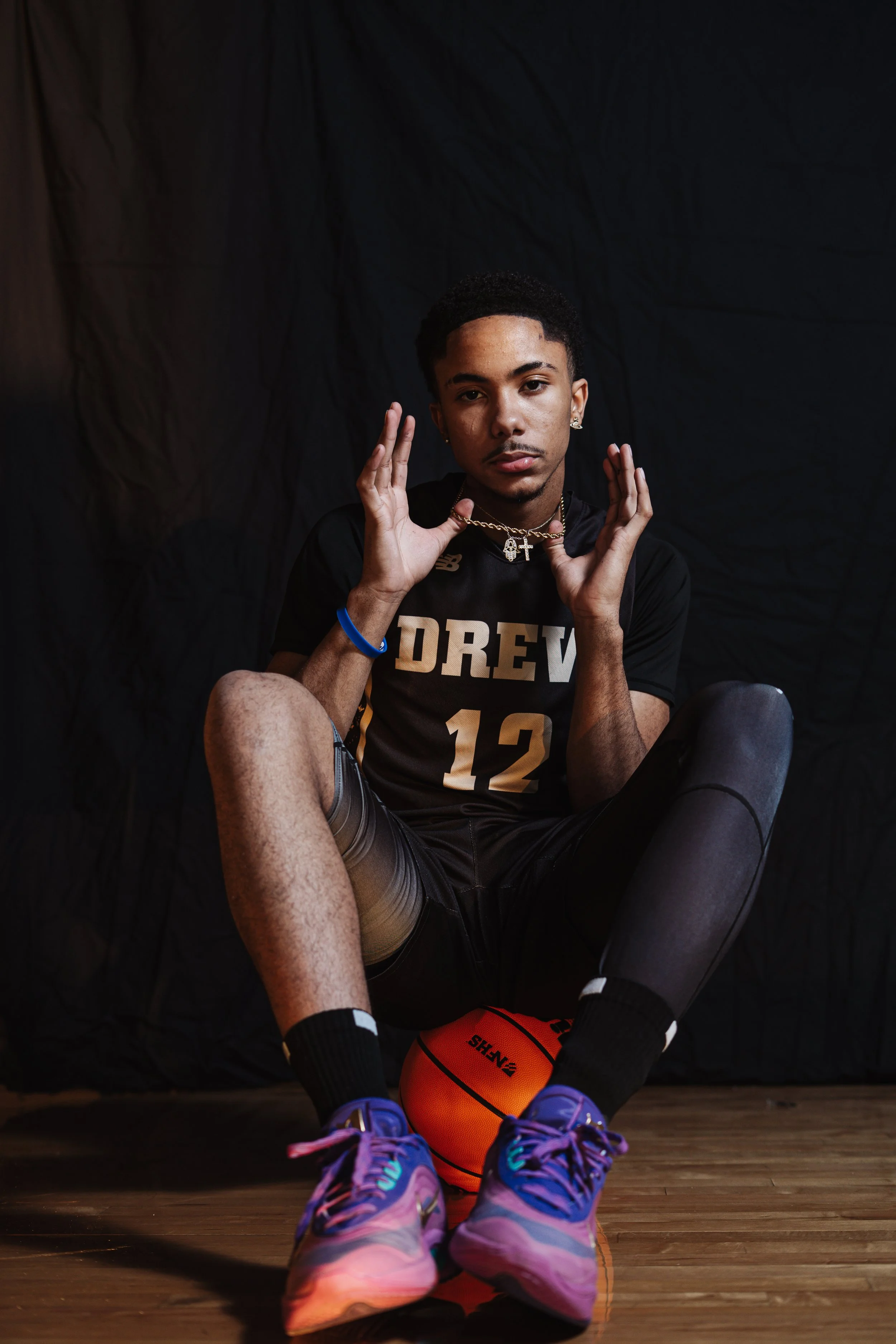 Young man sitting on a basketball court, holding a basketball between his feet, wearing a black sports jersey with the number 12, colorful sneakers, earrings, and necklaces, against a dark backdrop.