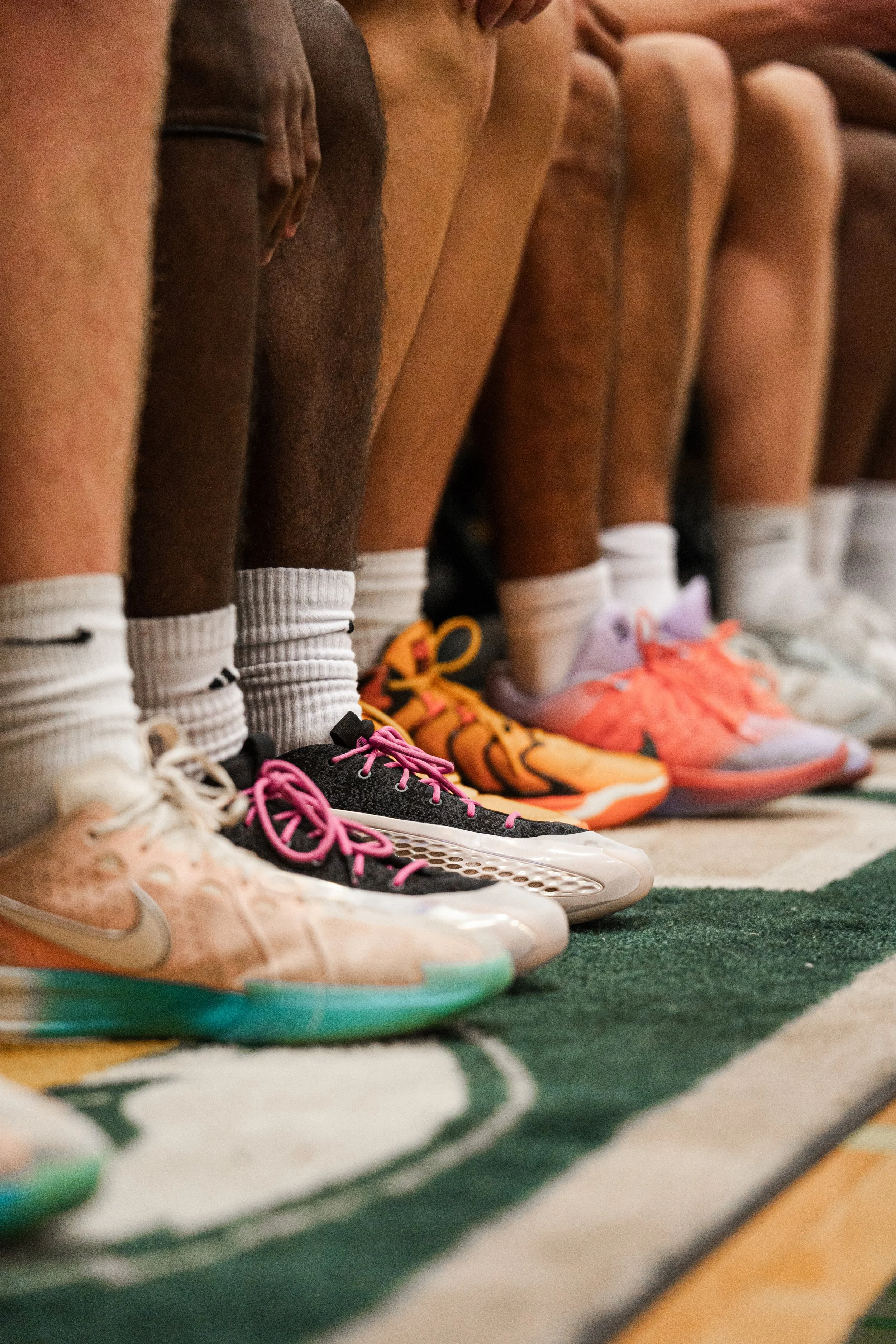 A lineup of basketball players' legs and feet, showing various colorful sneakers, standing on a basketball court.