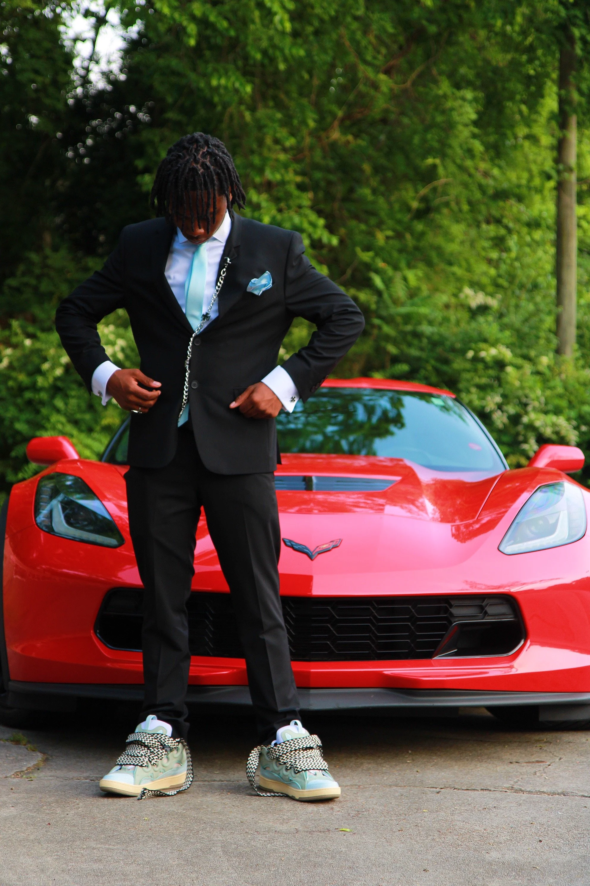 A man dressed in a black suit with a light blue tie and pocket square, wearing a chain, standing in front of a red sports car with a Chevrolet logo, on a driveway with green trees in the background.