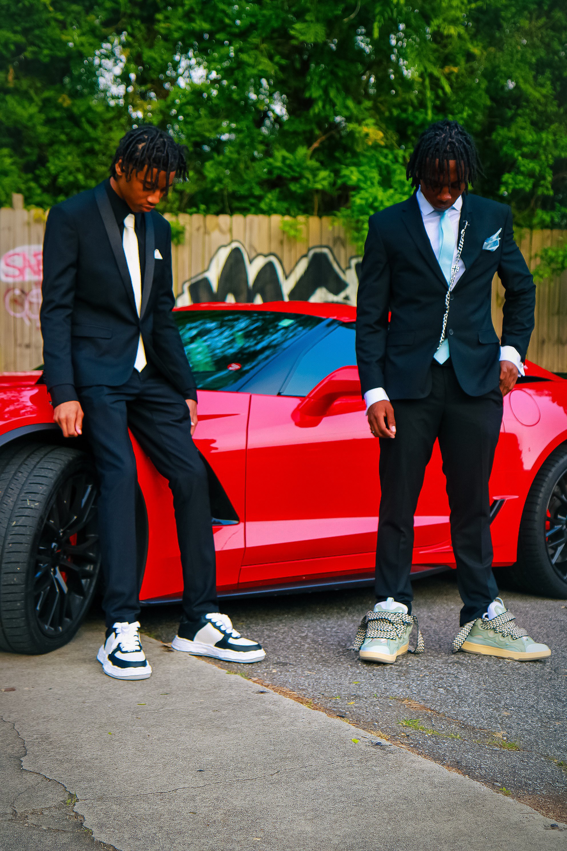 Two young men in suits standing beside a red sports car outside, with a wooden fence and green trees in the background.