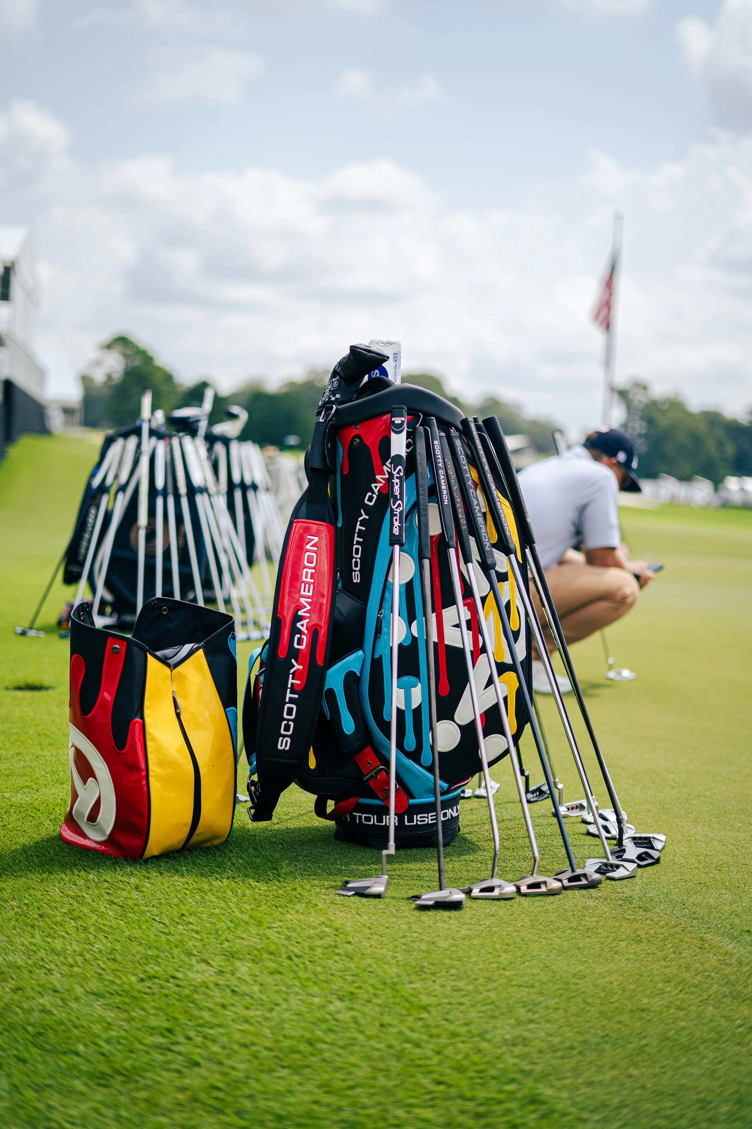 Golf clubs and bags arranged on a golf course green with a person squatting in the background, and an American flag in the distance.
