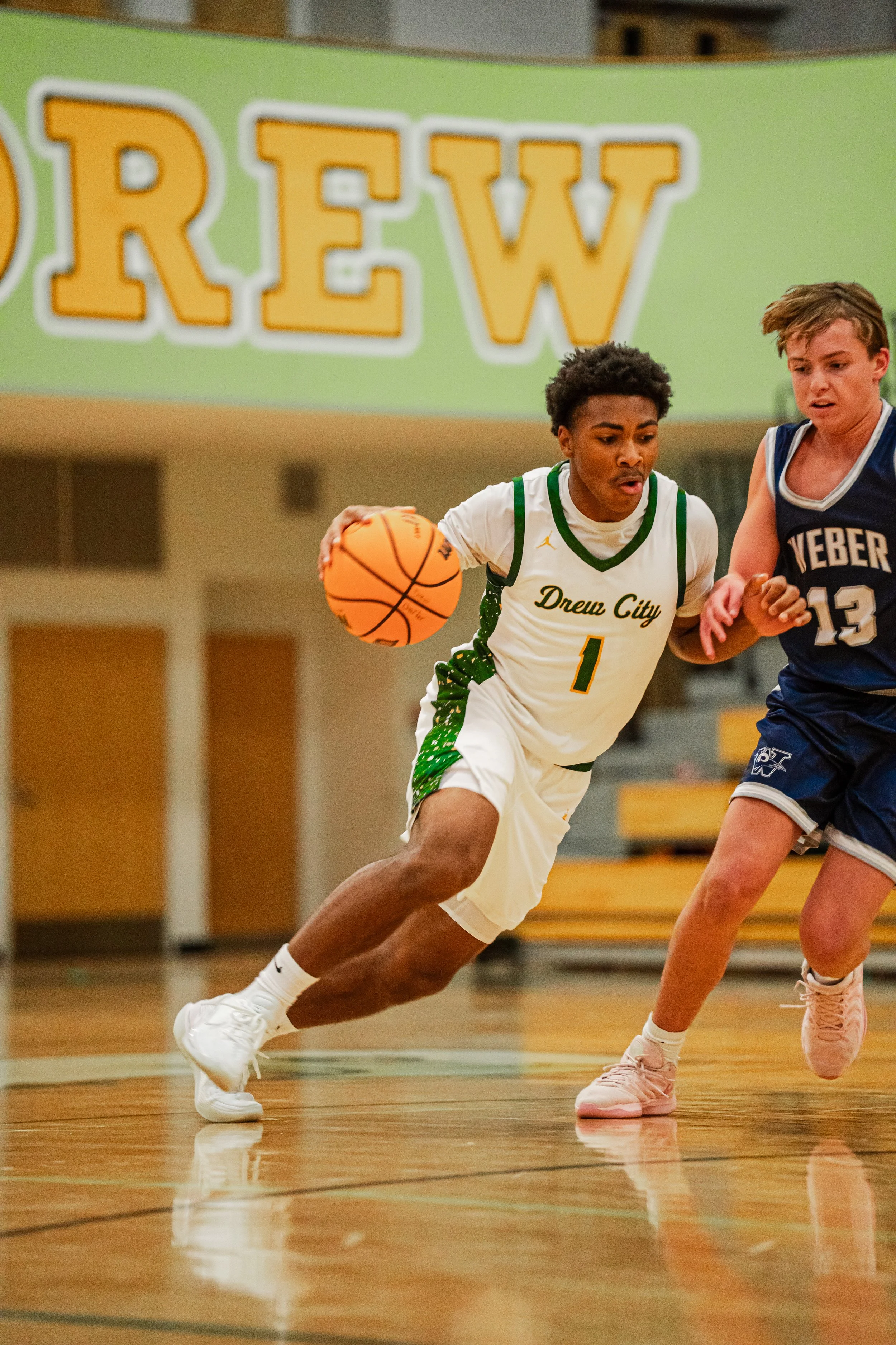 Two young male basketball players competing for the ball on an indoor court; one wears a white jersey with green accents, and the other wears a navy blue jersey. A large partial sign reading "REW" is visible in the background.