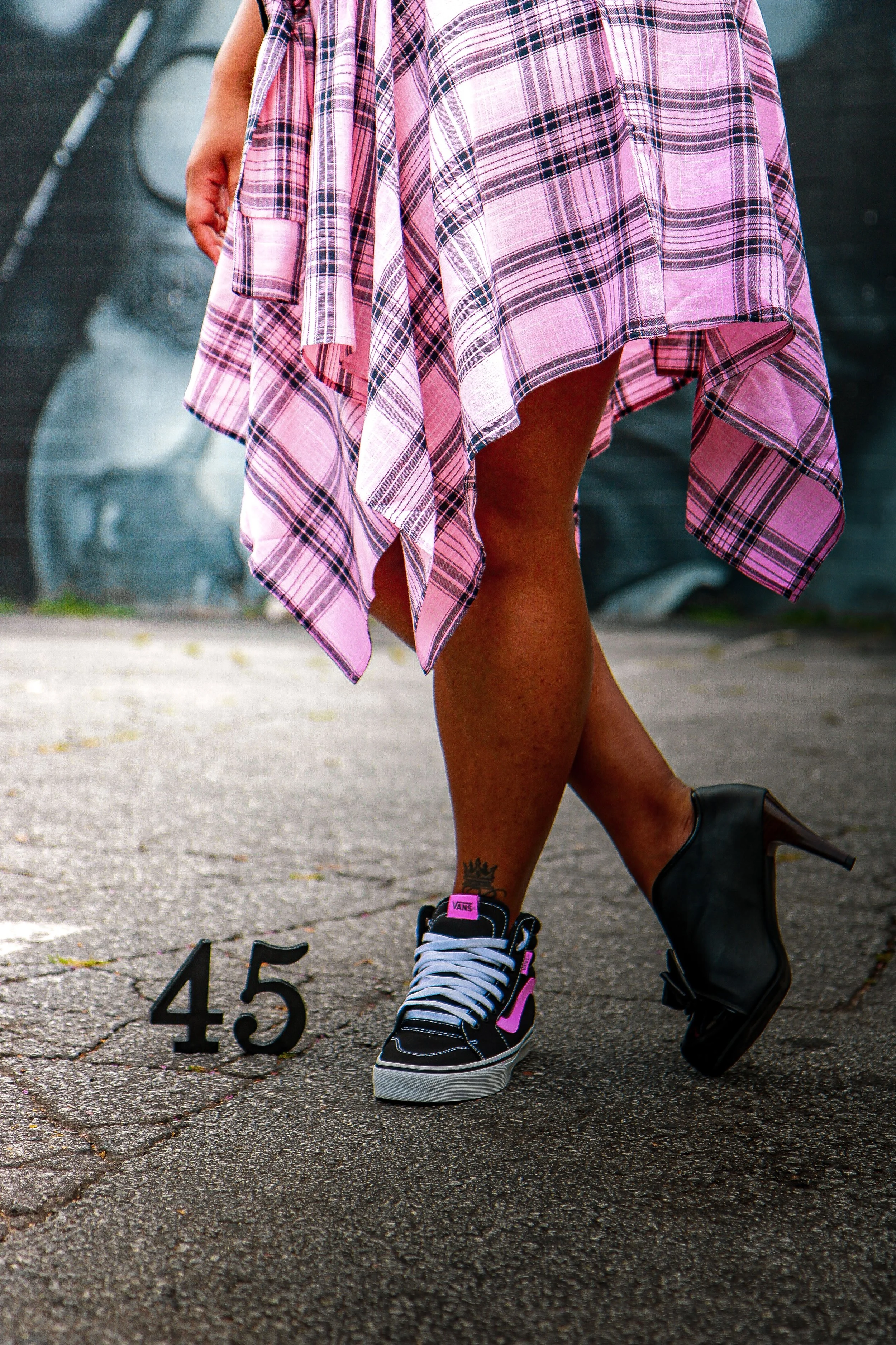 A person wearing a pink plaid dress with one leg extended, wearing black Vans sneakers with pink accents, standing next to black number markers '4' and '5' on an outdoor pavement.