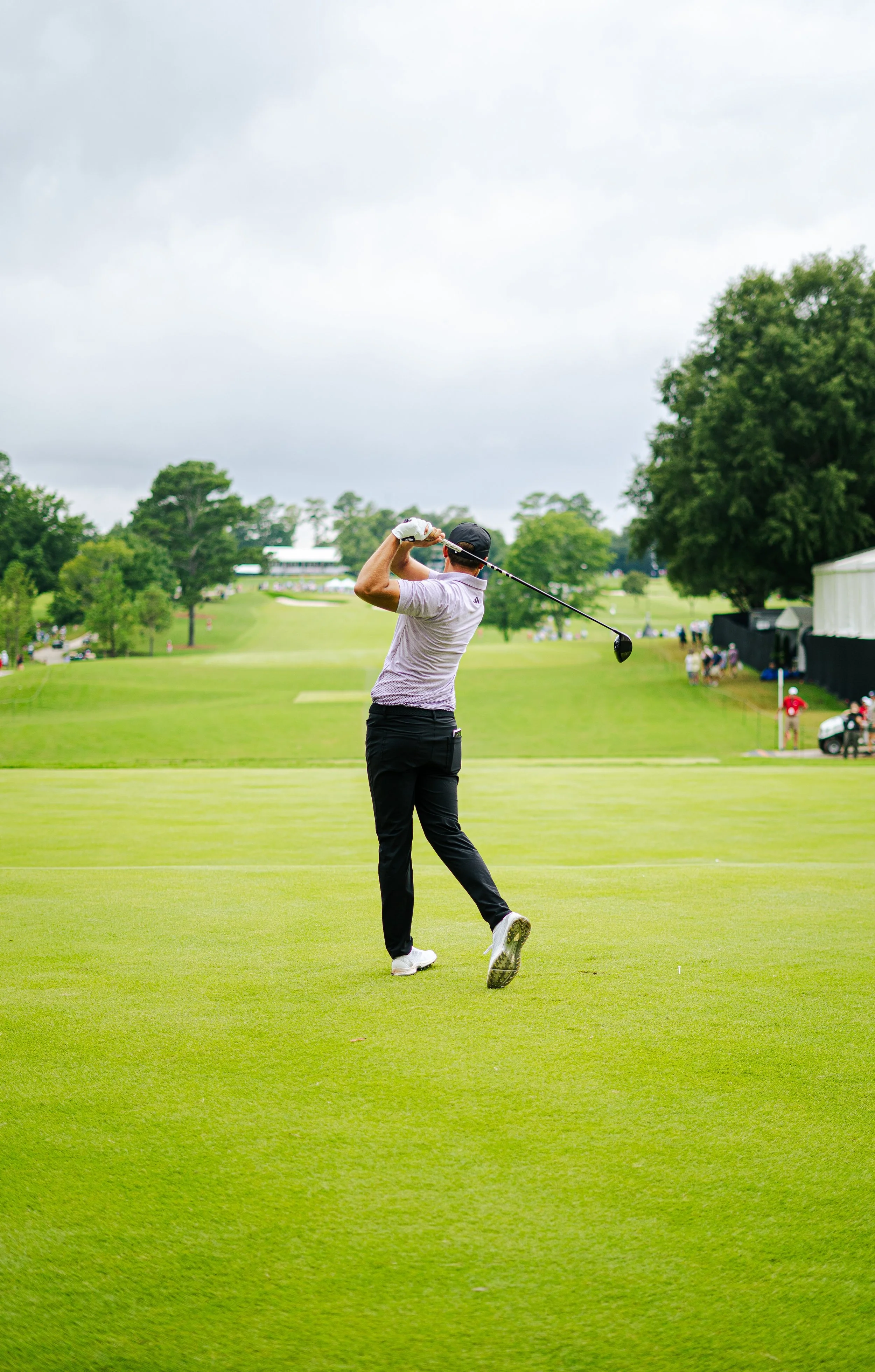 A man in a golf course swinging a golf club. The scene is outdoors with lush green grass and trees, and overcast sky.