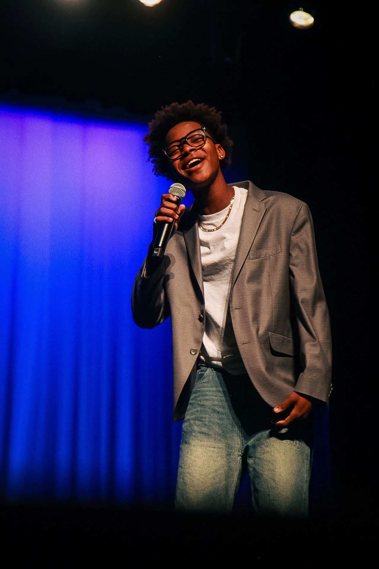 A person with glasses, wearing a gray blazer, white T-shirt, and jeans, is smiling and holding a microphone on stage with a blue curtain background.