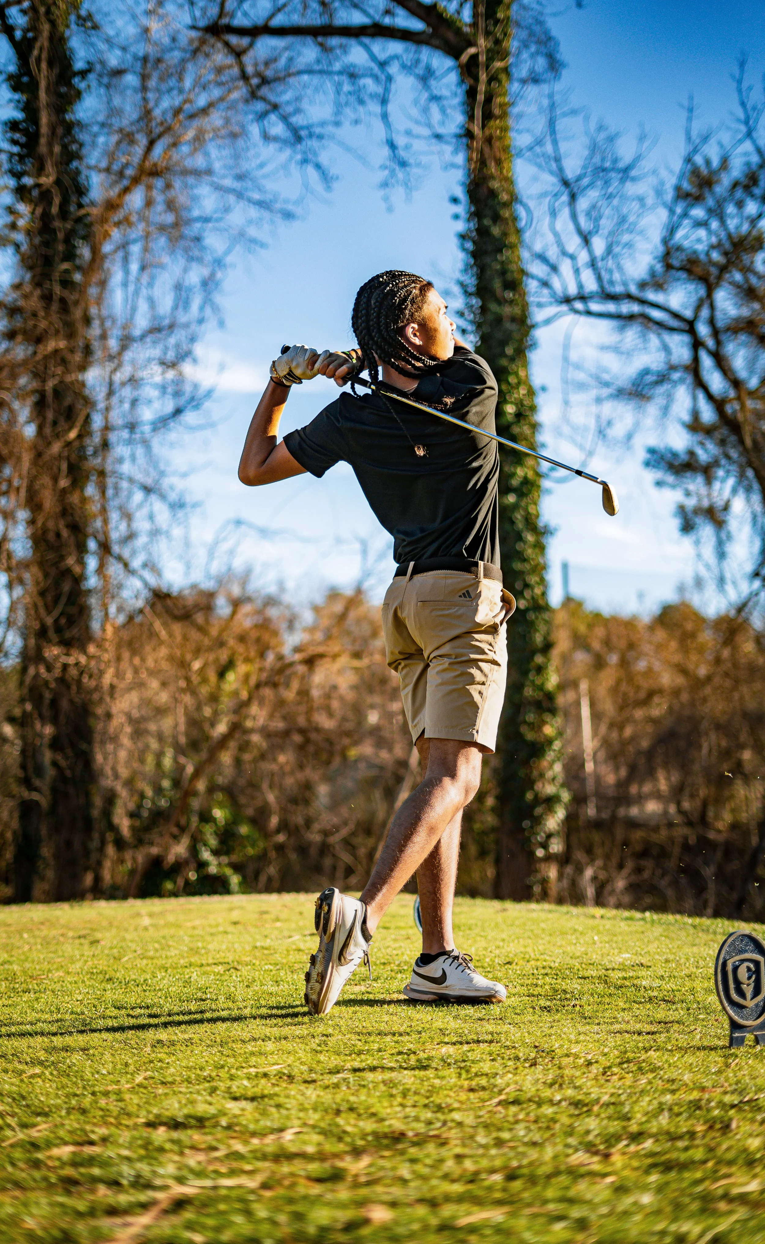 A person playing golf on a sunny day, mid-swing, on a golf course surrounded by trees.