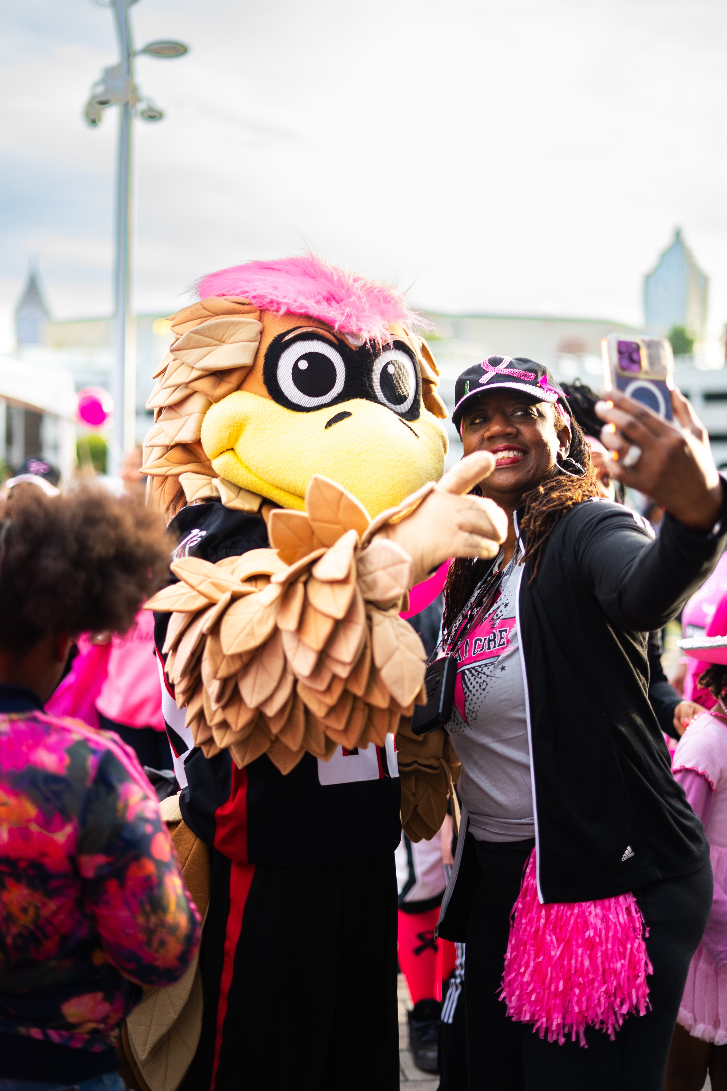 A person in a bird mascot costume with pink hair knuckles with a woman taking a selfie at an outdoor event. People dressed in pink are visible in the background.