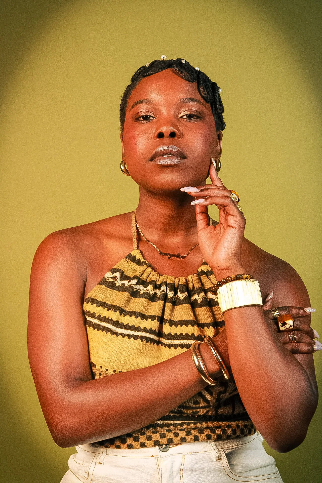Portrait of a woman with dark skin, wearing a yellow and black patterned top, gold jewelry, and with styled hair, posing against a yellow-green background.