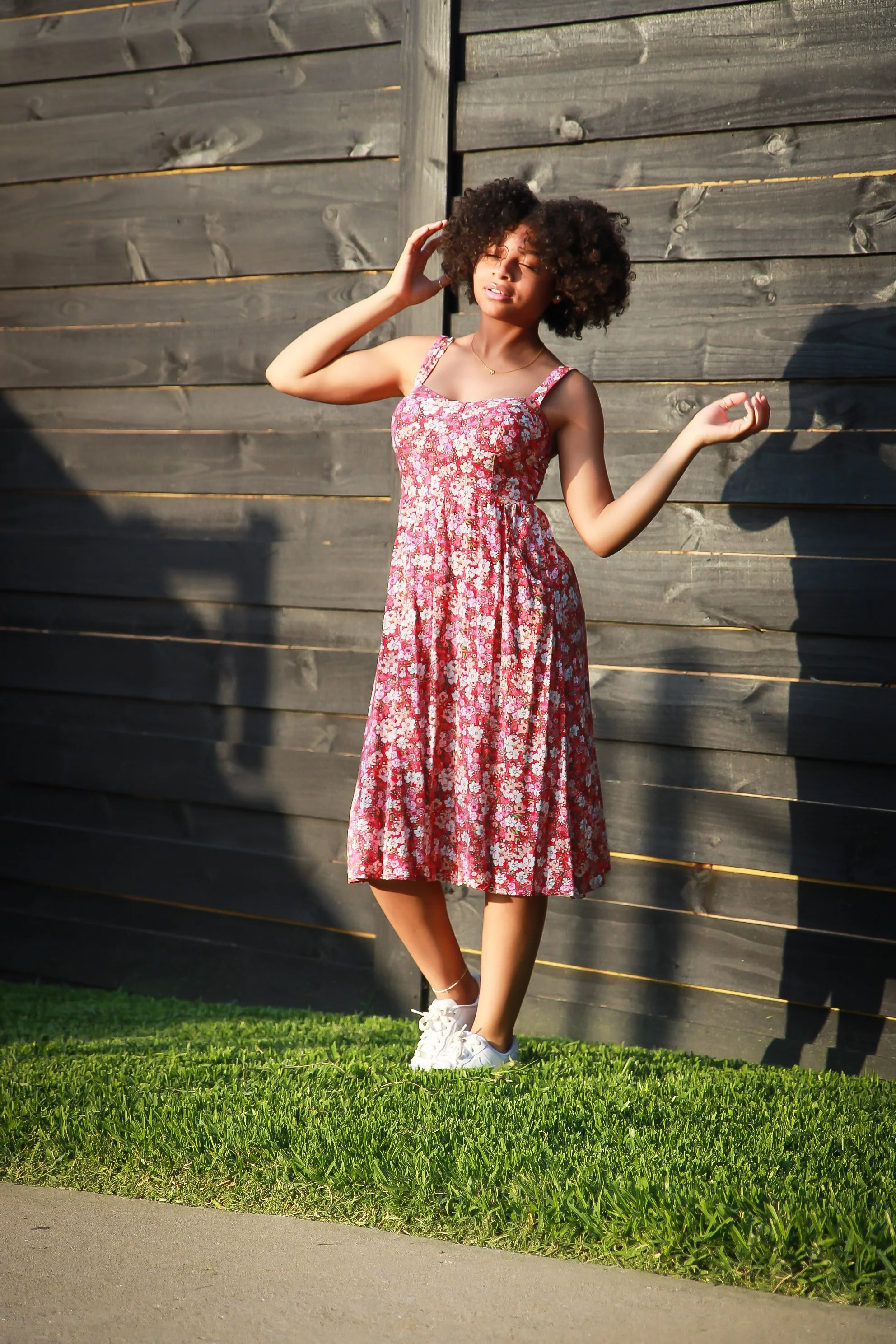 A woman with curly hair standing outdoors against a wooden fence, wearing a floral dress and sneakers, with her eyes closed and one hand in her hair.